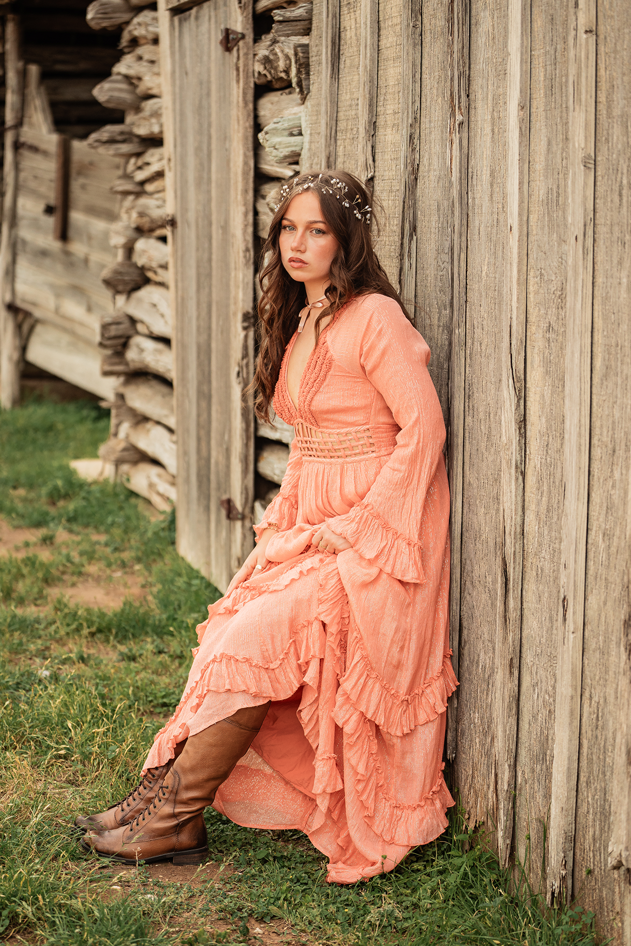 A young woman with long brown hair and a floral headband, wearing a peach-colored bohemian dress and brown lace-up boots, leaning against a rustic wooden barn wall.