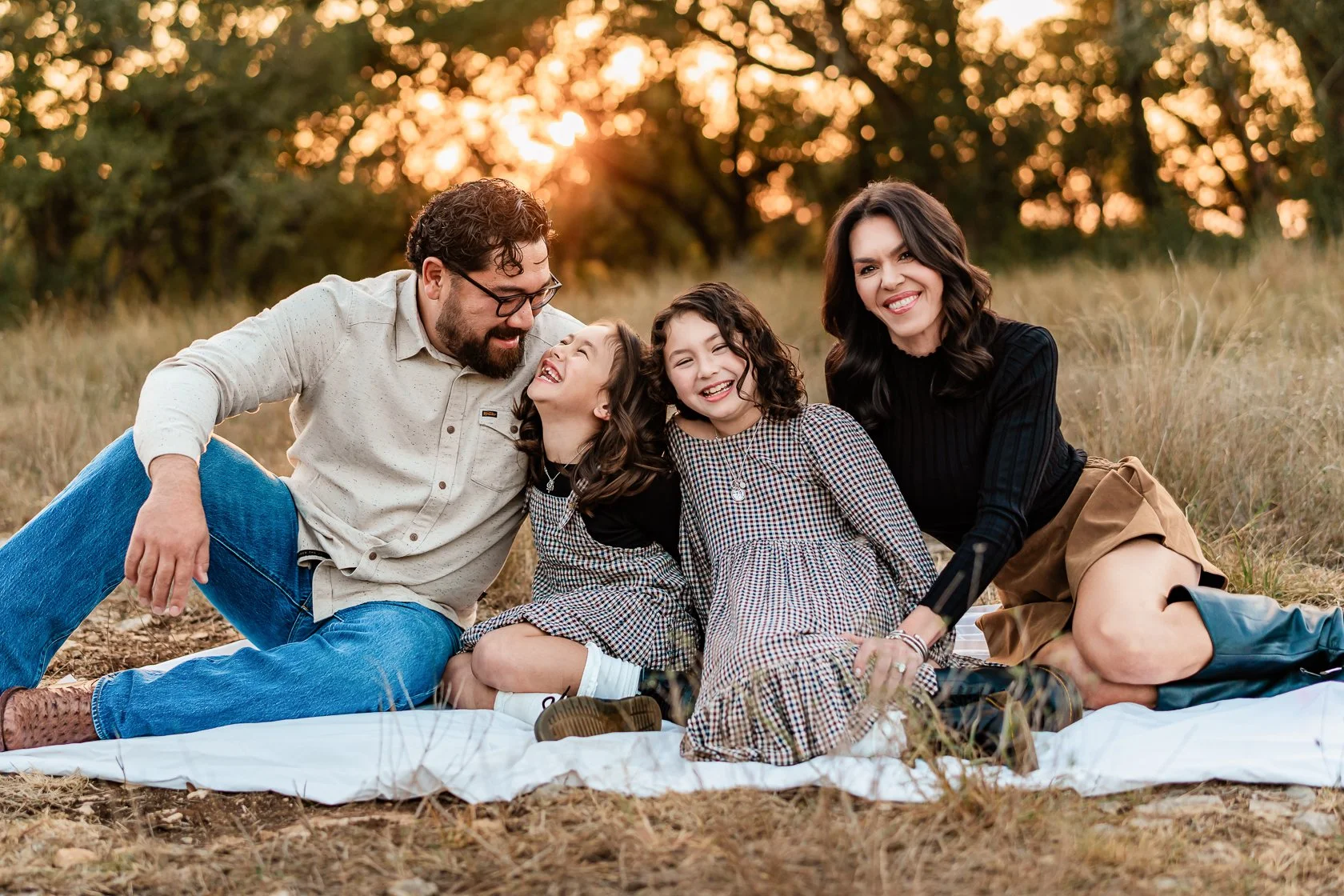 A family of four, two adults and two children, sitting on a white blanket outdoors during sunset, laughing and enjoying each other's company in a grassy field.