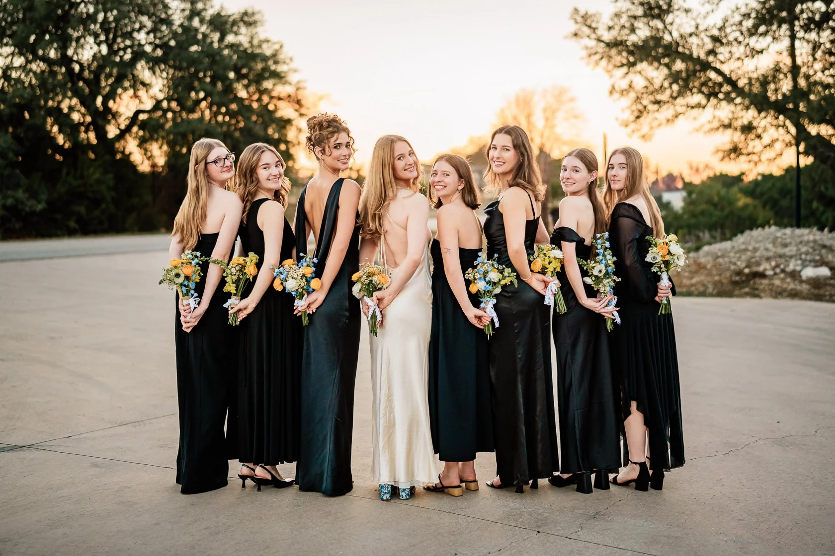 Group of women in black dresses holding bouquets of flowers outdoors during sunset