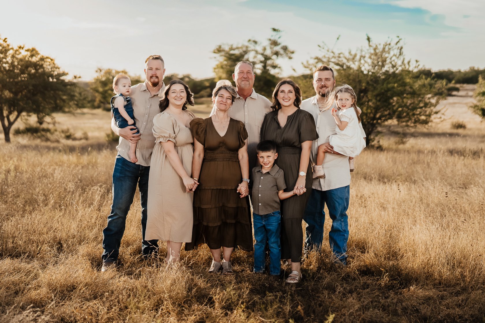 Family group portrait outdoors in a field during golden hour, consisting of adults and children holding hands and smiling.
