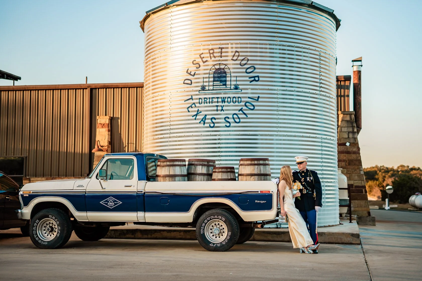 A newlywed couple standing next to a vintage pickup truck with barrels in the truck bed, in front of a large metal water tank with the words "Desert Door Texas Sunset" painted on it, during sunset at a winery or distillery.