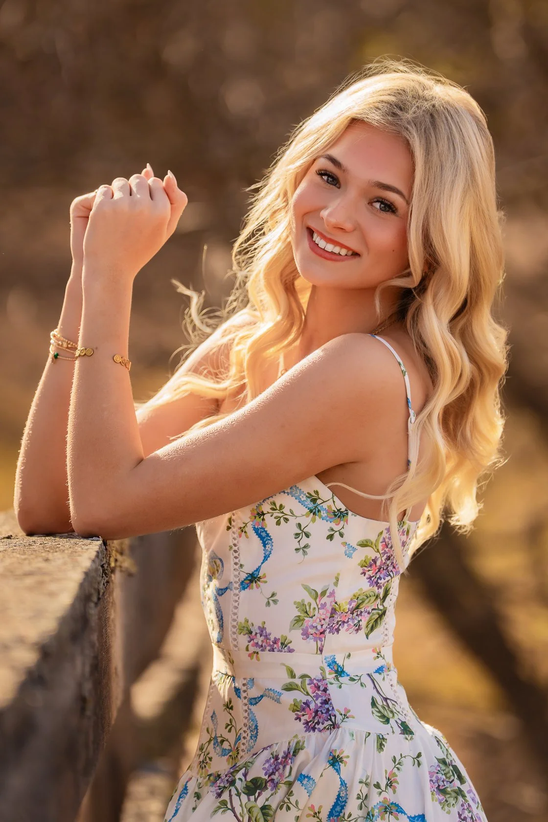 A young woman with long, wavy blonde hair smiling at the camera, leaning on a stone wall outdoors during golden hour, wearing a floral dress and jewelry.