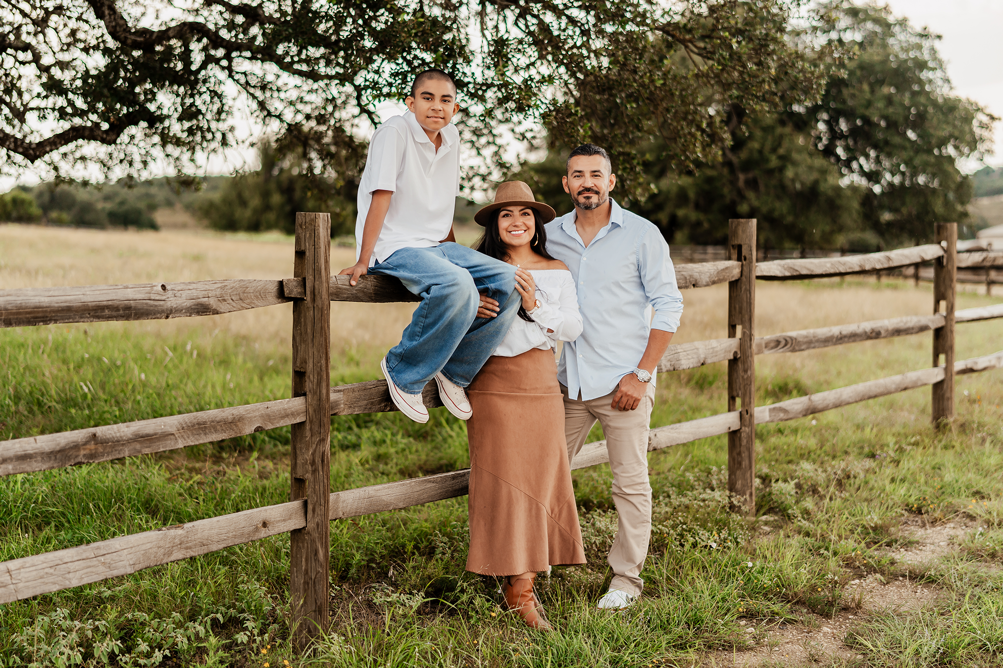 Family of four outdoor, standing in front of a wooden fence with green grass and trees, smiling at the camera.