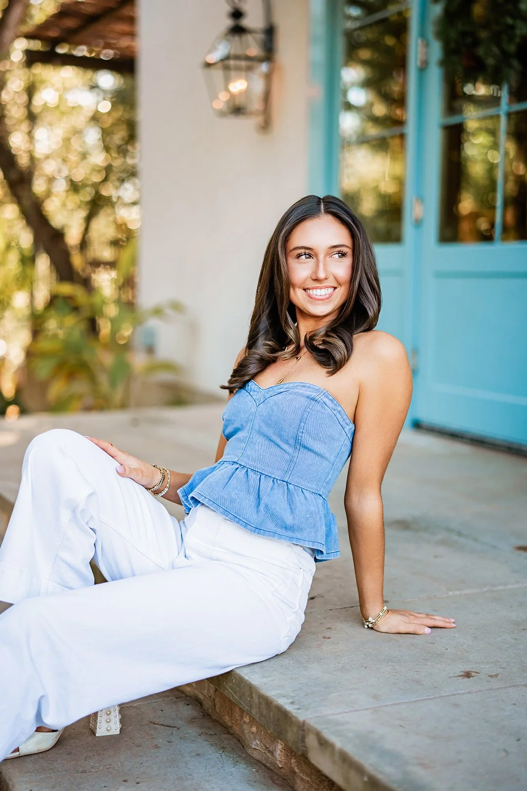 A young woman with long dark hair sitting on a concrete porch, smiling and looking to her right. She is wearing a strapless blue top and white pants, with jewelry on her wrist and neck. The background includes a light-colored wall, a turquoise door, and some greenery.