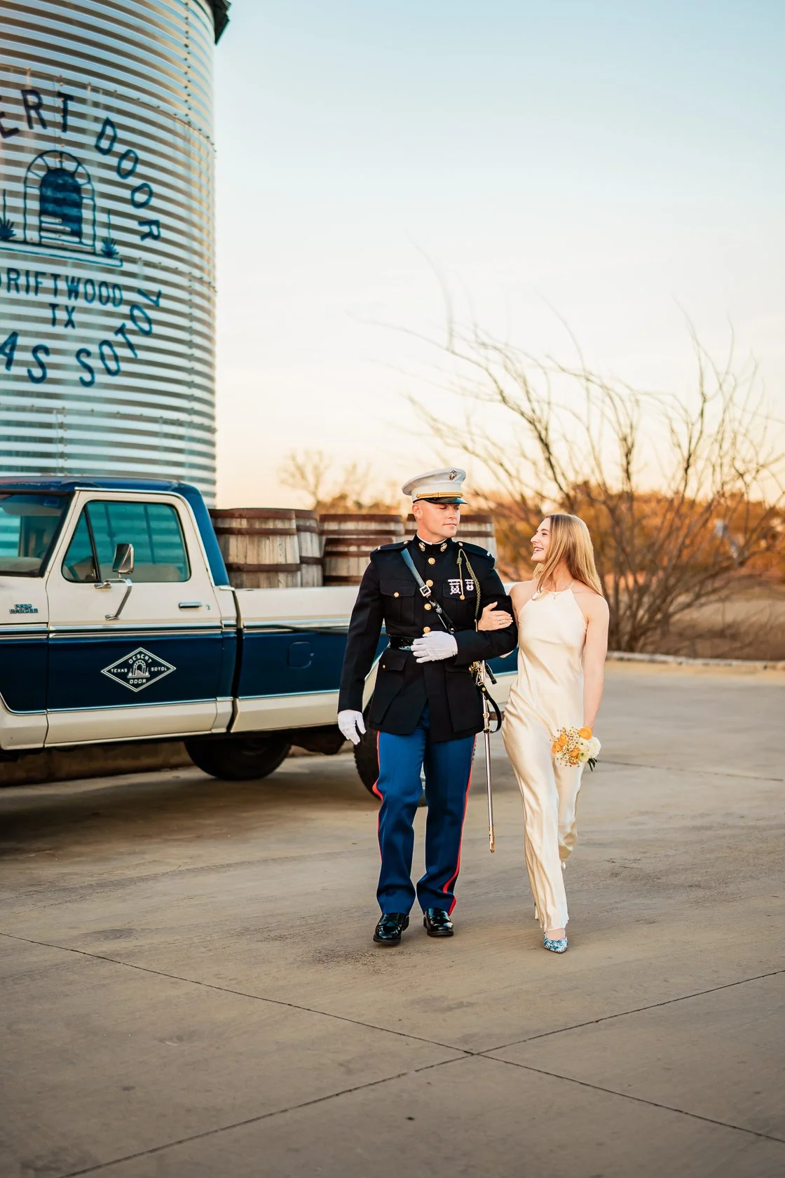 A man in a military uniform walking arm-in-arm with a woman in a white dress holding a small bouquet, near a vintage blue and white pickup truck and a large silos in the background during sunset.