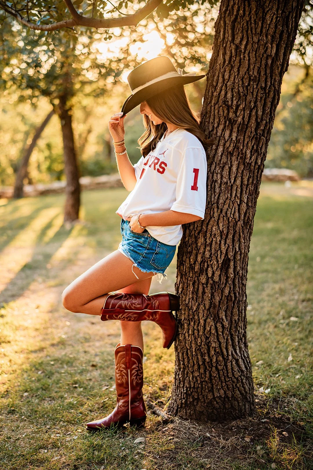 A young woman leaning against a tree in a park during sunset, wearing a black cowboy hat, white sports jersey, denim shorts, and tall brown cowboy boots.