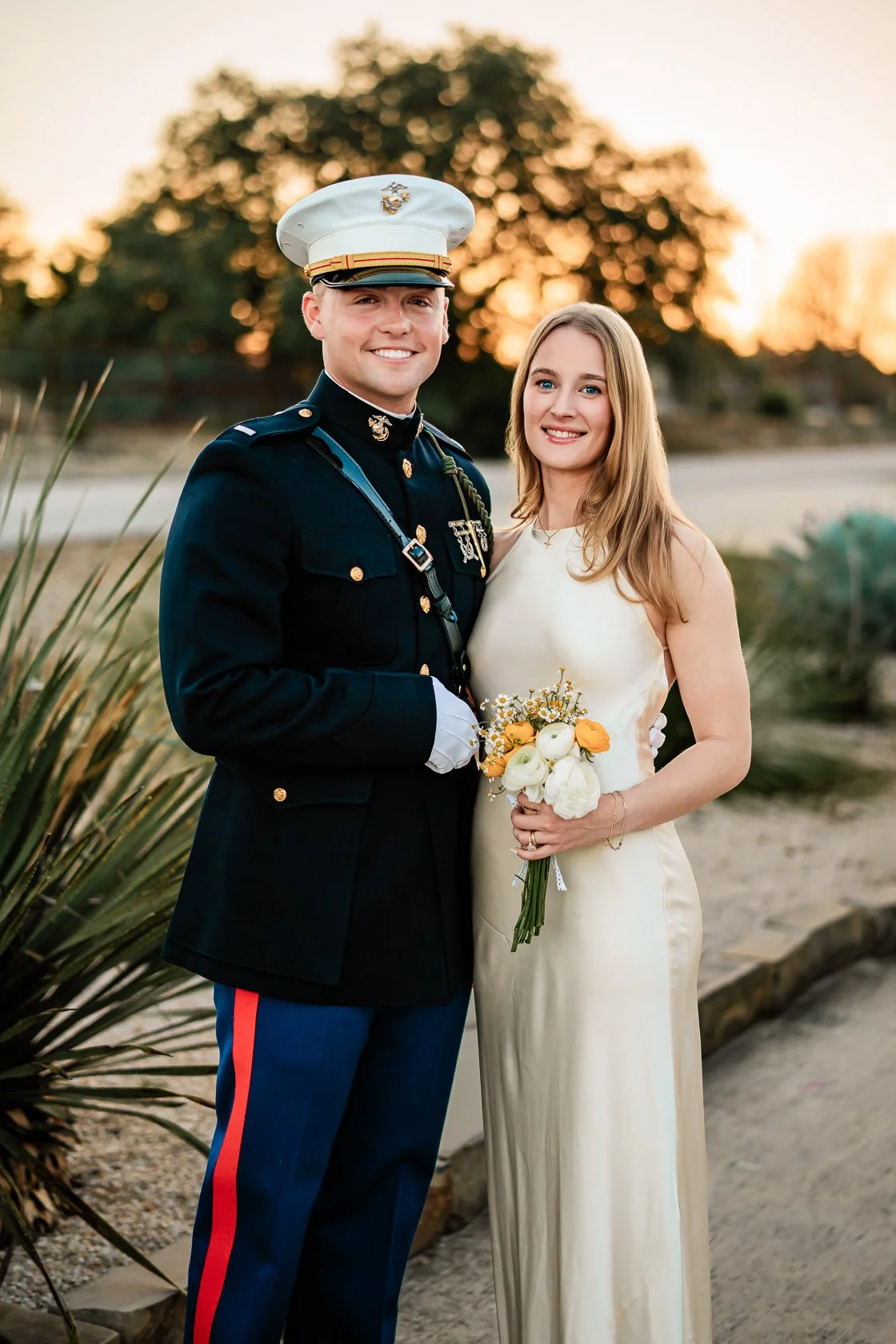 A couple dressed in wedding attire, with one person in a military uniform and the other in a white dress, standing outdoors during sunset, holding a bouquet of flowers.