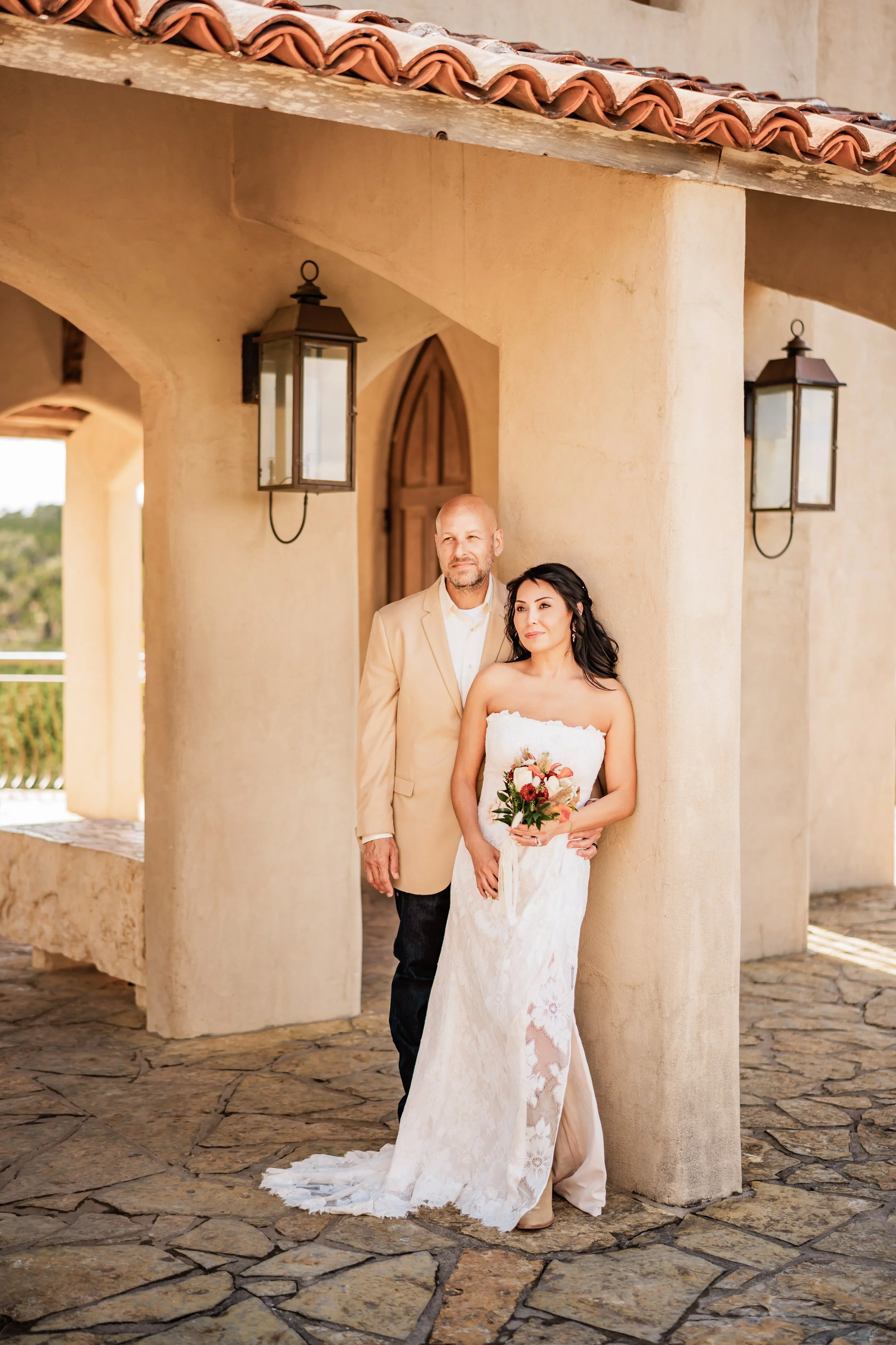 A bride in a strapless white wedding dress holding a bouquet stands next to a man in a beige blazer and white shirt, outdoors near a Mediterranean style building with beige stucco walls, wooden door, and black lanterns mounted on the wall.
