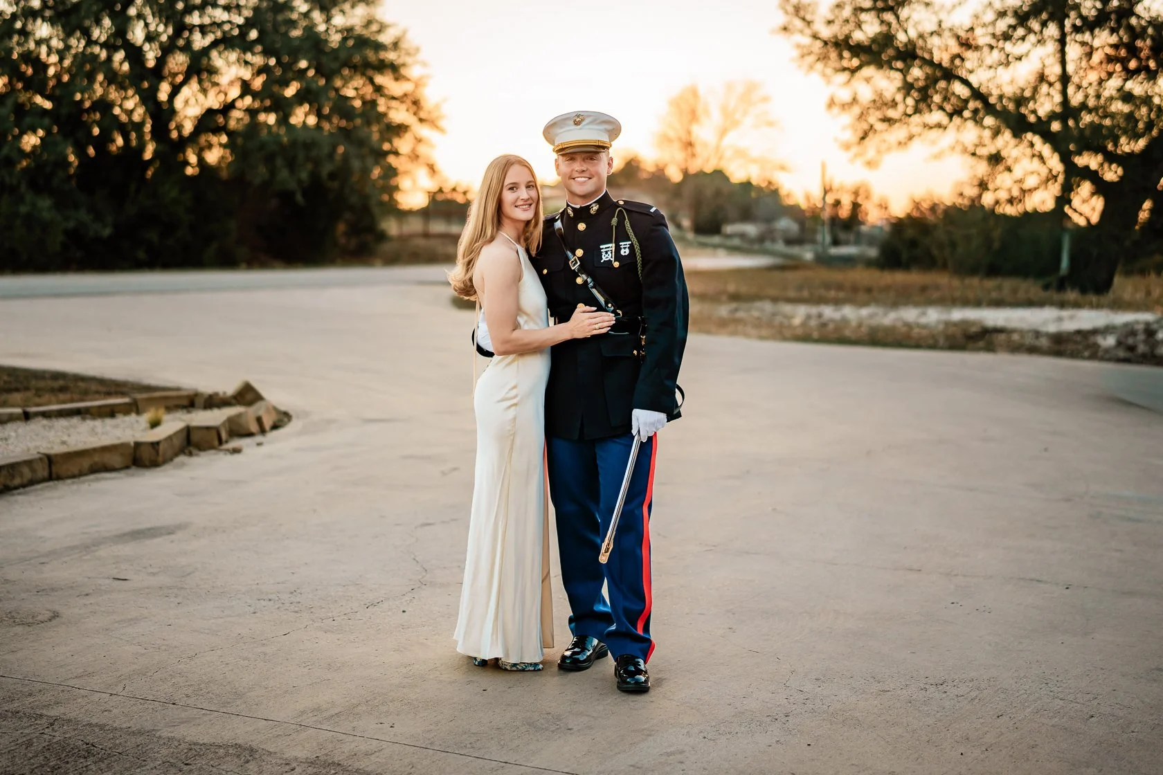 A young woman and a man in U.S. Marine dress uniform posing together outdoors at sunset.
