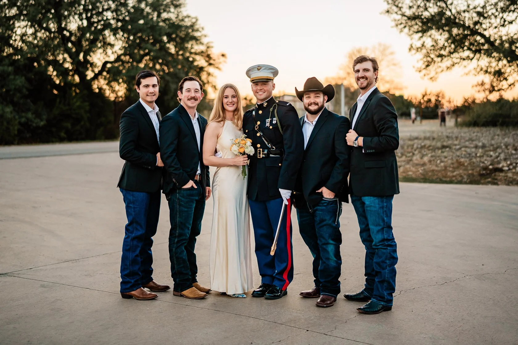 Group of six people, including a woman in a light-colored dress holding a bouquet, and a man in a military uniform, outdoors during sunset.