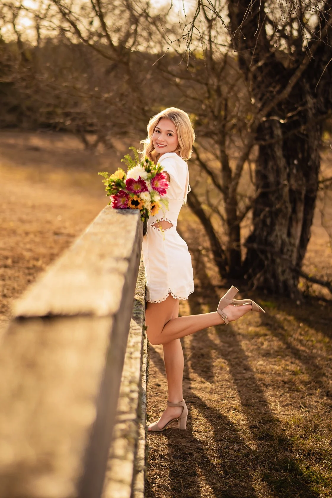 A woman in a white dress and nude heels leaning against a wooden fence in a park during sunset, holding a bouquet of colorful flowers and smiling.