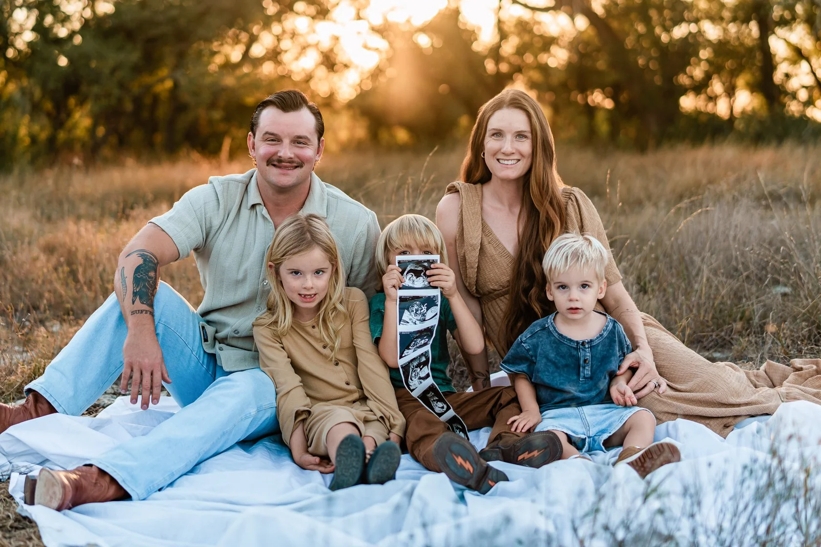 A family of five sitting on a blanket outdoors during sunset, with two parents and three children, one of whom is holding ultrasound pictures.
