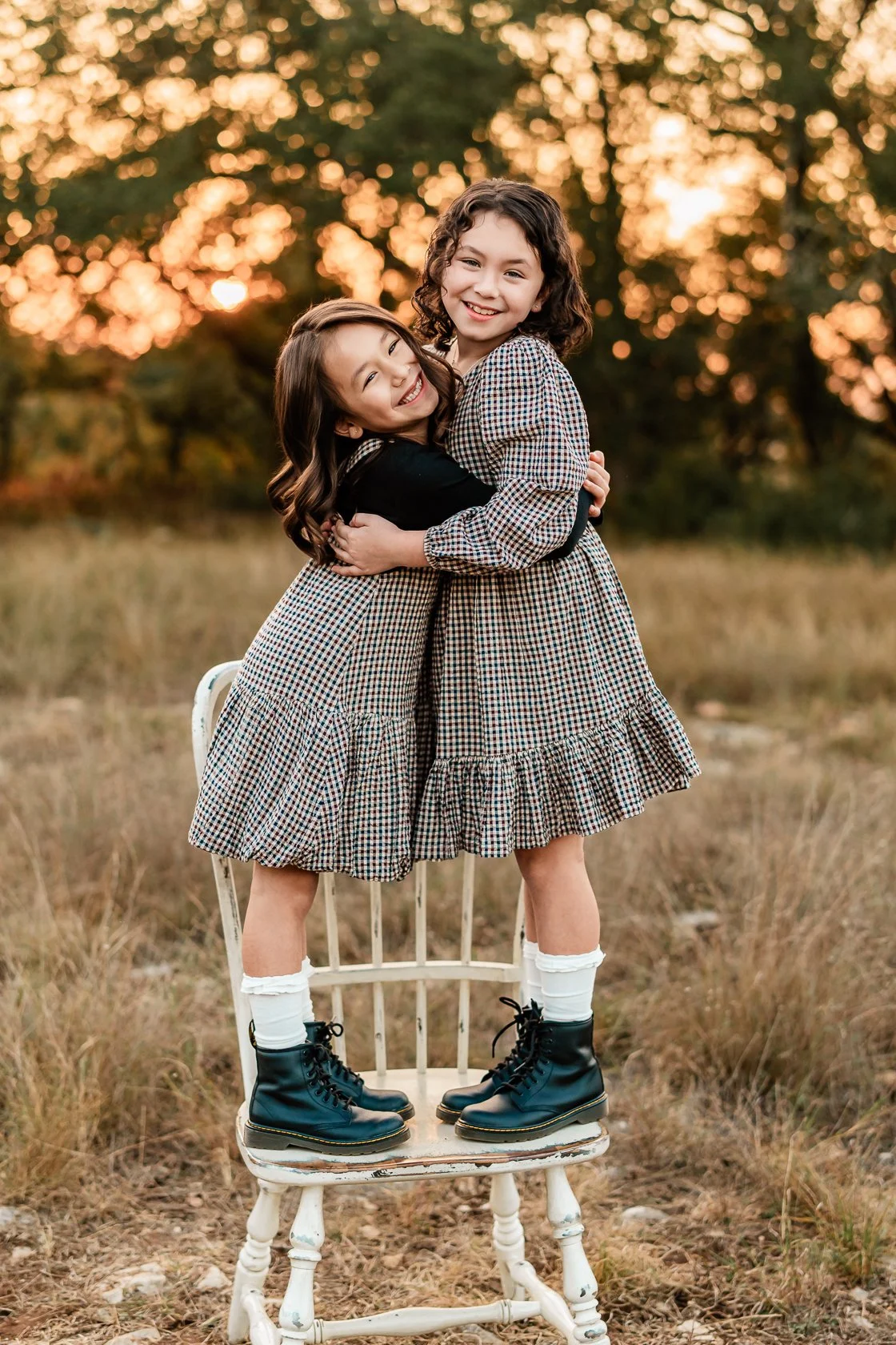 Two young girls in matching plaid dresses hugging each other and smiling outdoors during sunset. One girl stands on a white wooden chair, balancing on her tiptoes in black boots and white socks, with a background of trees and orange sunset sky.