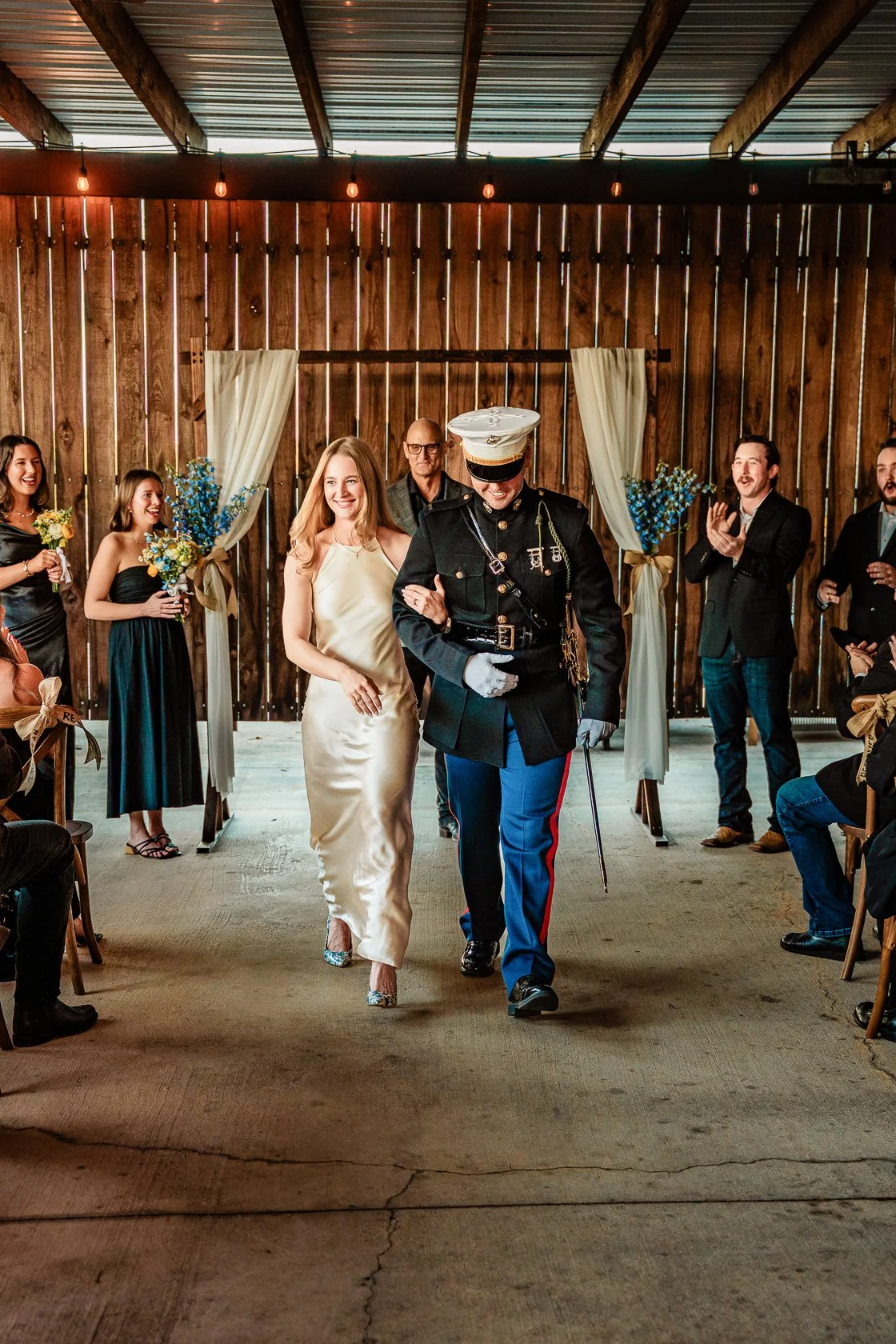 A wedding reception with a bride walking down the aisle arm-in-arm with a military officer in uniform. Guests are smiling and clapping in a rustic venue with wooden walls and string lights.