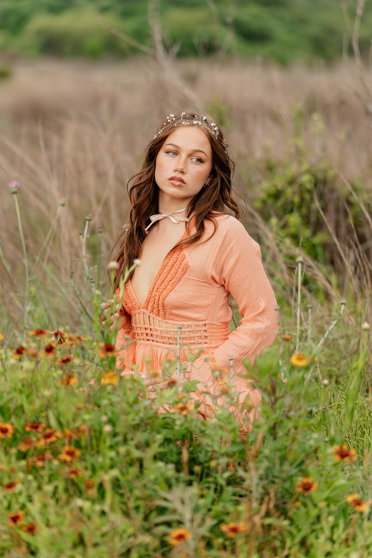 A young woman with long brunette hair wearing a peach-colored dress and a pearl headband stands in a field of wildflowers and grass, looking to her right with a contemplative expression.