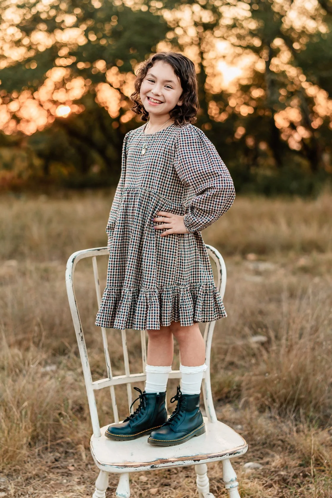 Young girl standing on a white vintage chair outdoors during sunset, smiling and posing with hand on her hip, wearing a plaid dress, white socks, and black lace-up boots.
