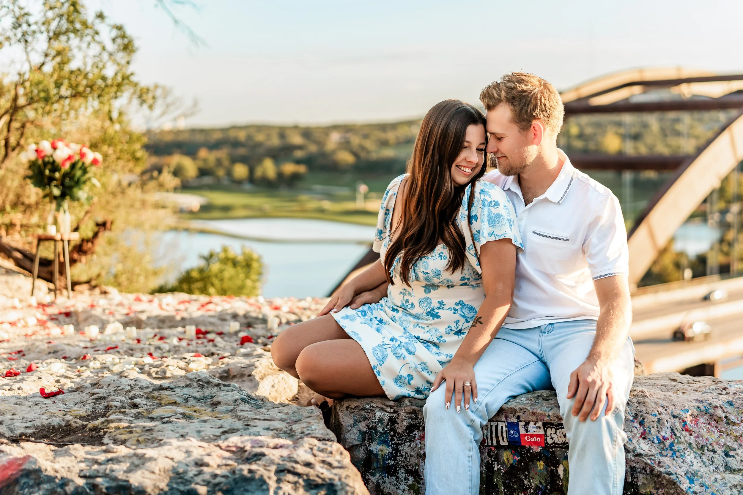 A young couple sitting on a stone ledge with a view of a river and bridge in the background. They are smiling and leaning close to each other on a sunny day.