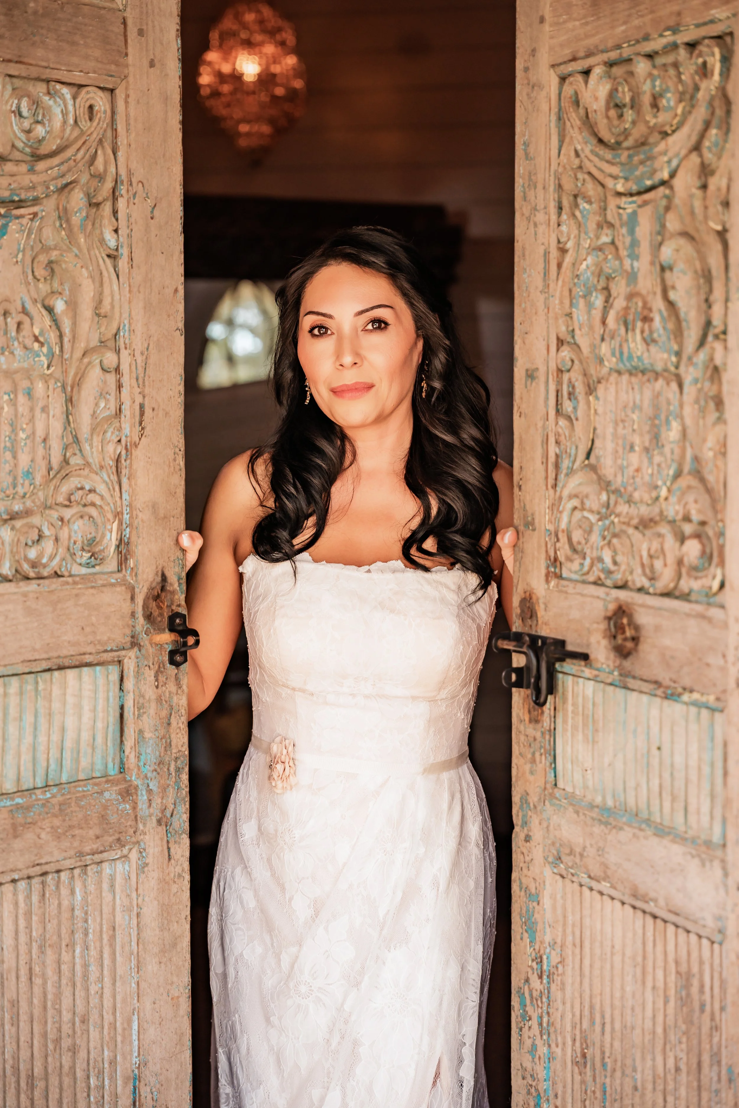 A woman with long dark hair, wearing a white lace dress, standing in a rustic doorway with ornate carved wood and opening slightly, looking directly at the camera.