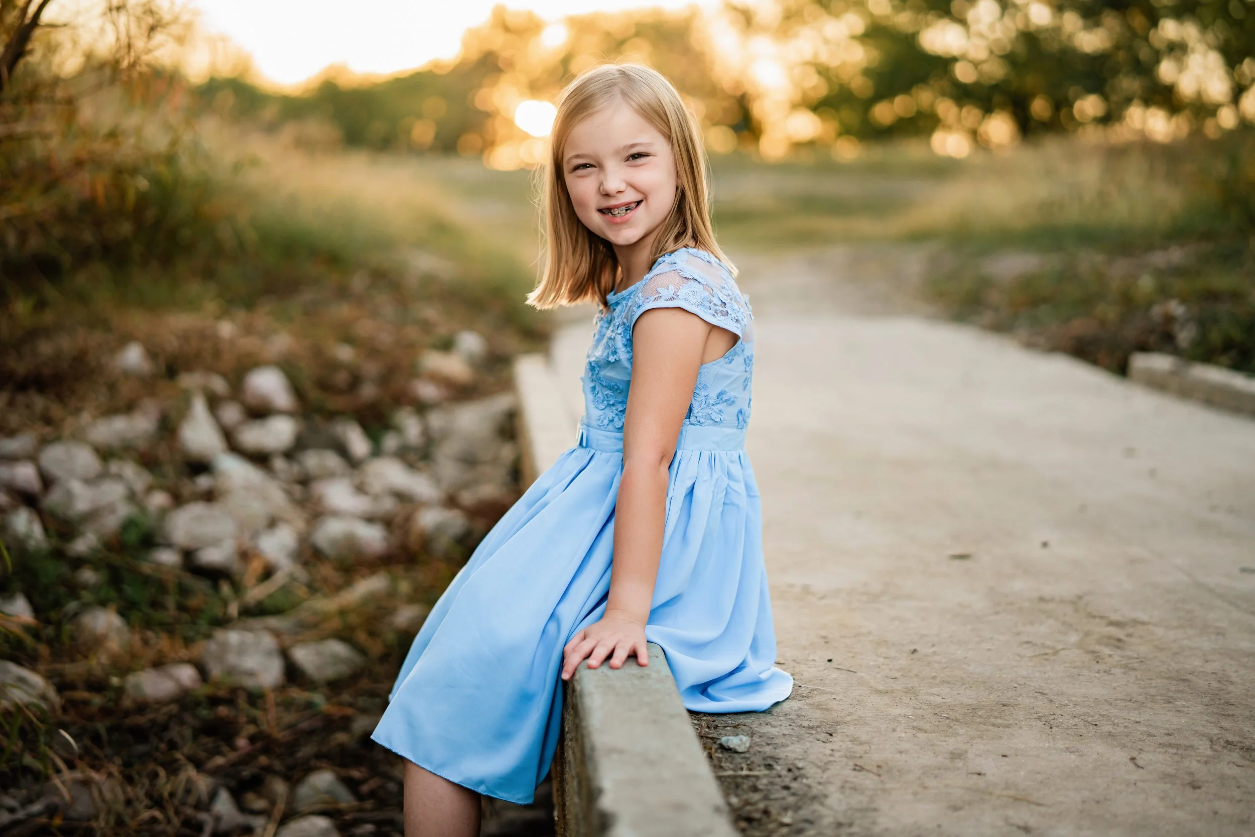 A young girl in a light blue dress sitting on a low concrete barrier outdoors during sunset, smiling at the camera.