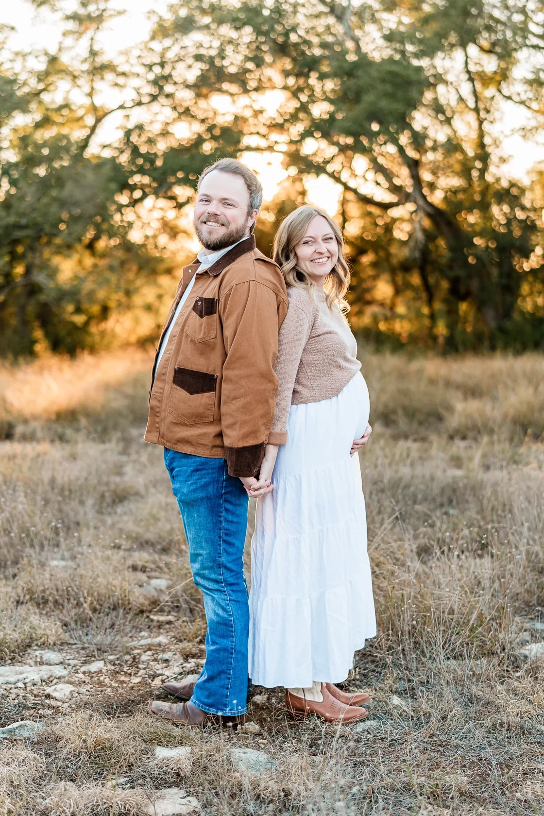 A couple stands outdoors in a field during sunset, holding hands, with the pregnant woman smiling and the man smiling at the camera.