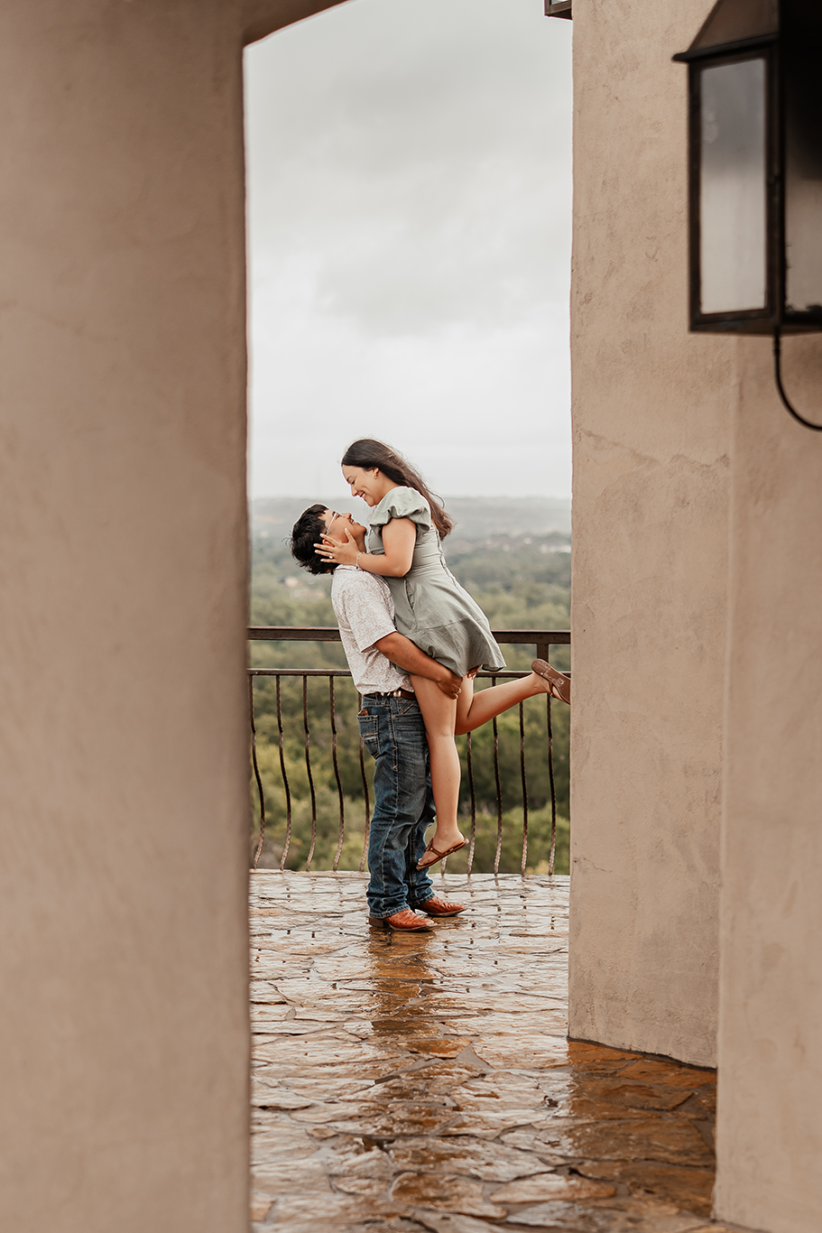 A man lifting a woman in his arms on a balcony, both smiling and looking at each other, with a scenic landscape in the background on a cloudy day.
