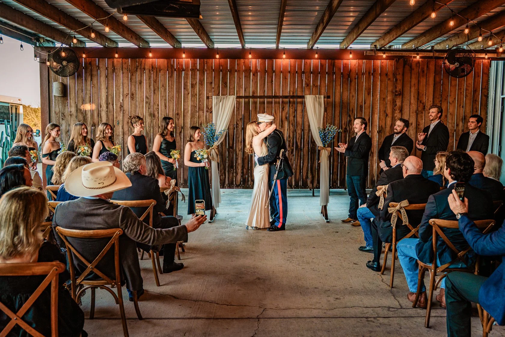 A wedding ceremony with a bride and groom kissing at the altar. Bridesmaids and groomsmen are standing on either side, and guests are seated on wooden chairs, some taking photos. The setting is rustic with wood paneling and string lights overhead.