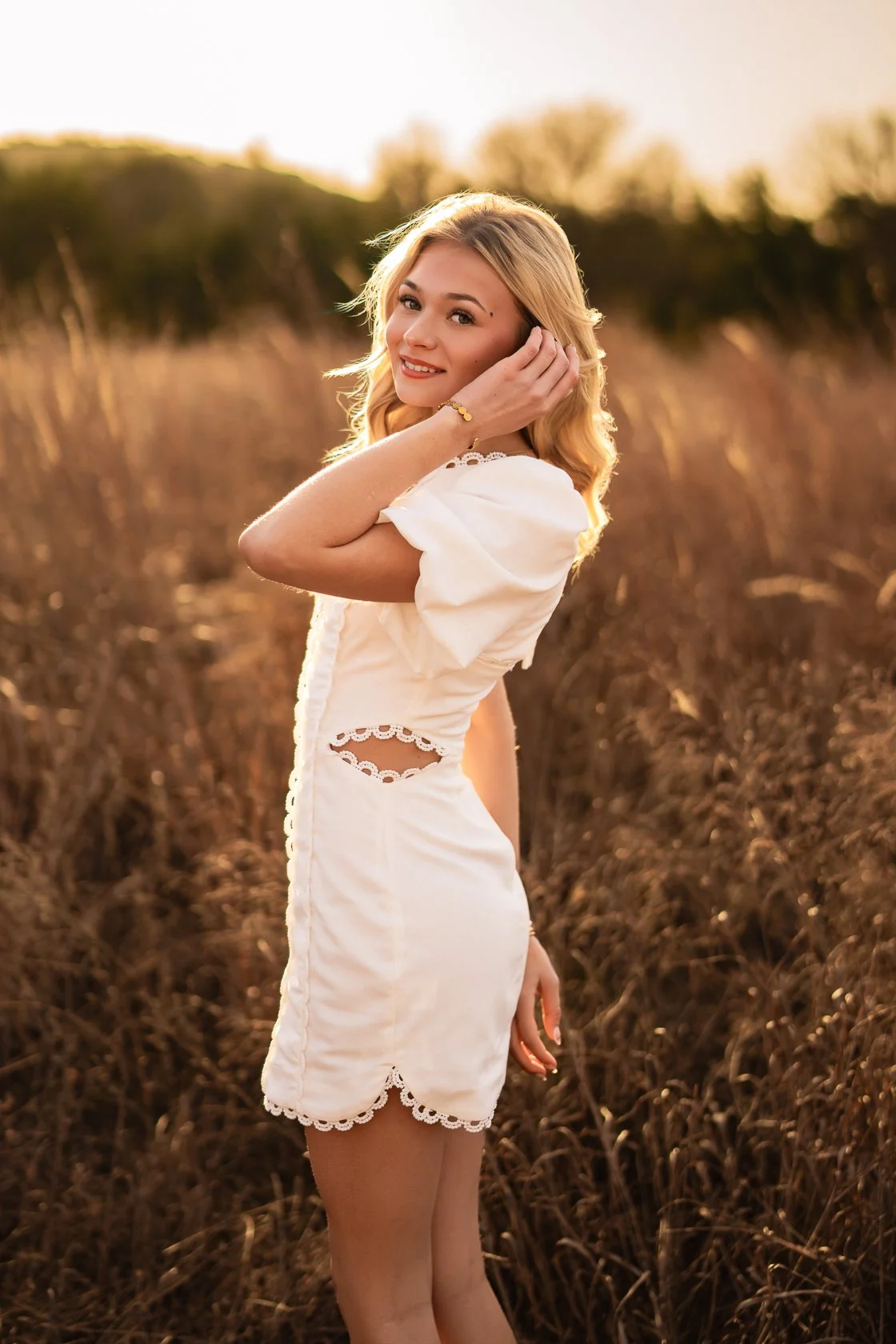 A young woman in a white dress standing in a field during sunset, smiling and touching her hair.