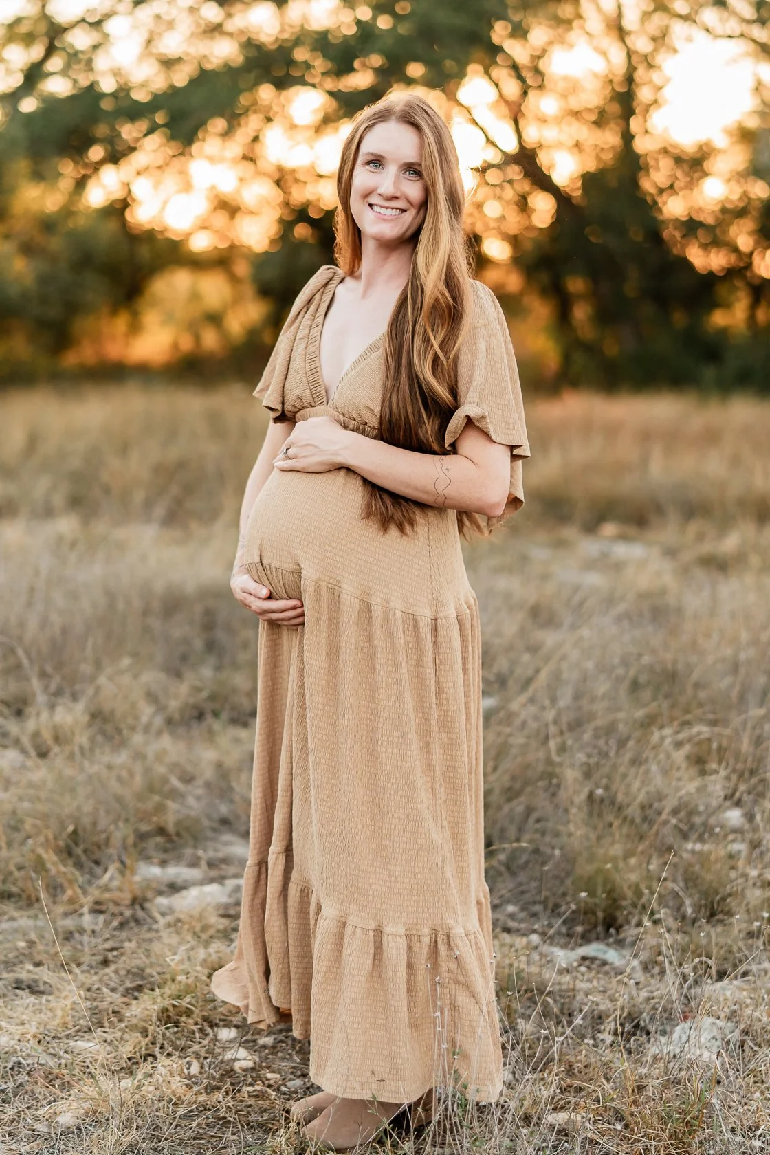 A pregnant woman with long red hair in a tan dress standing outdoors at sunset, smiling and holding her belly.