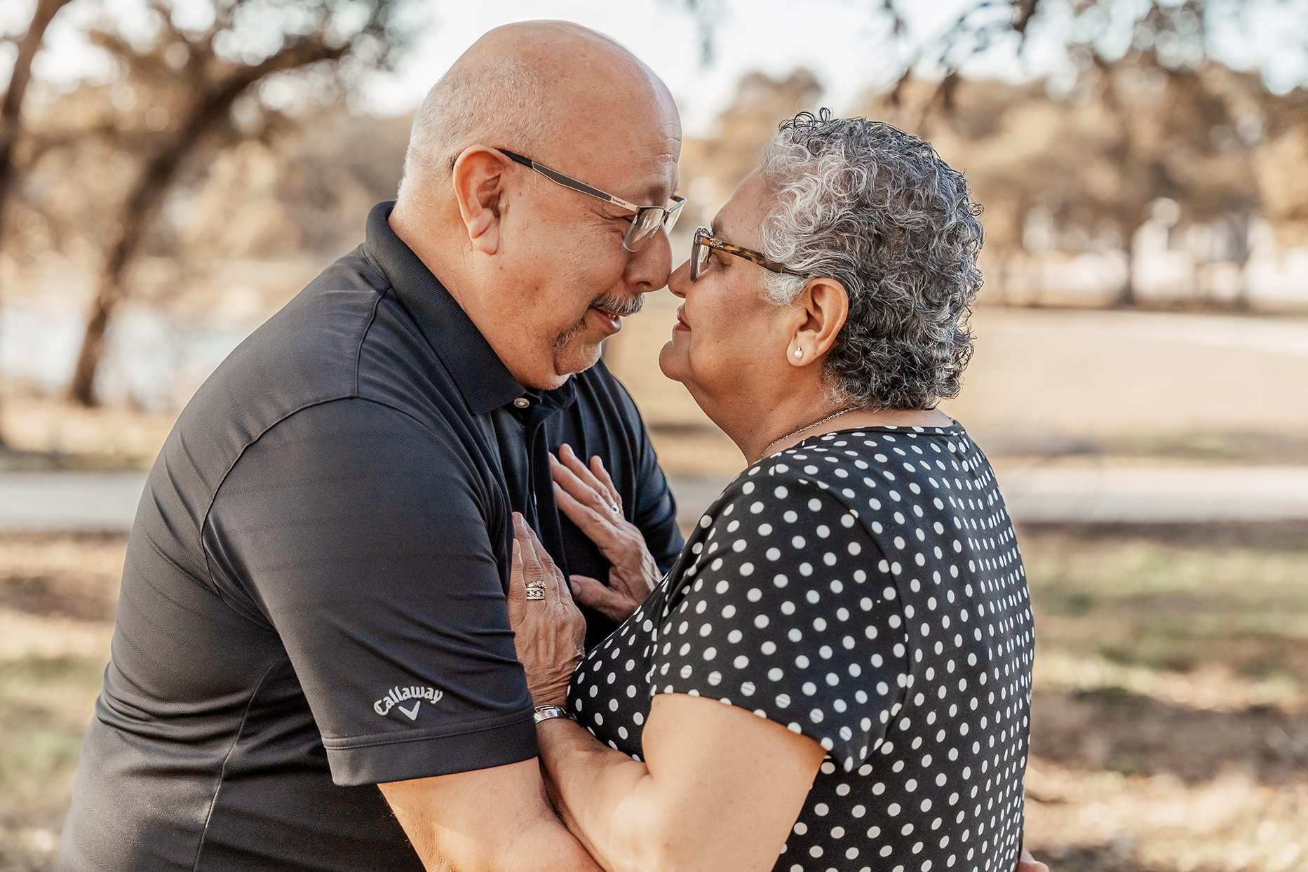 An elderly couple standing outdoors in a park, touching foreheads and smiling at each other, with the woman holding her hands on the man's chest.