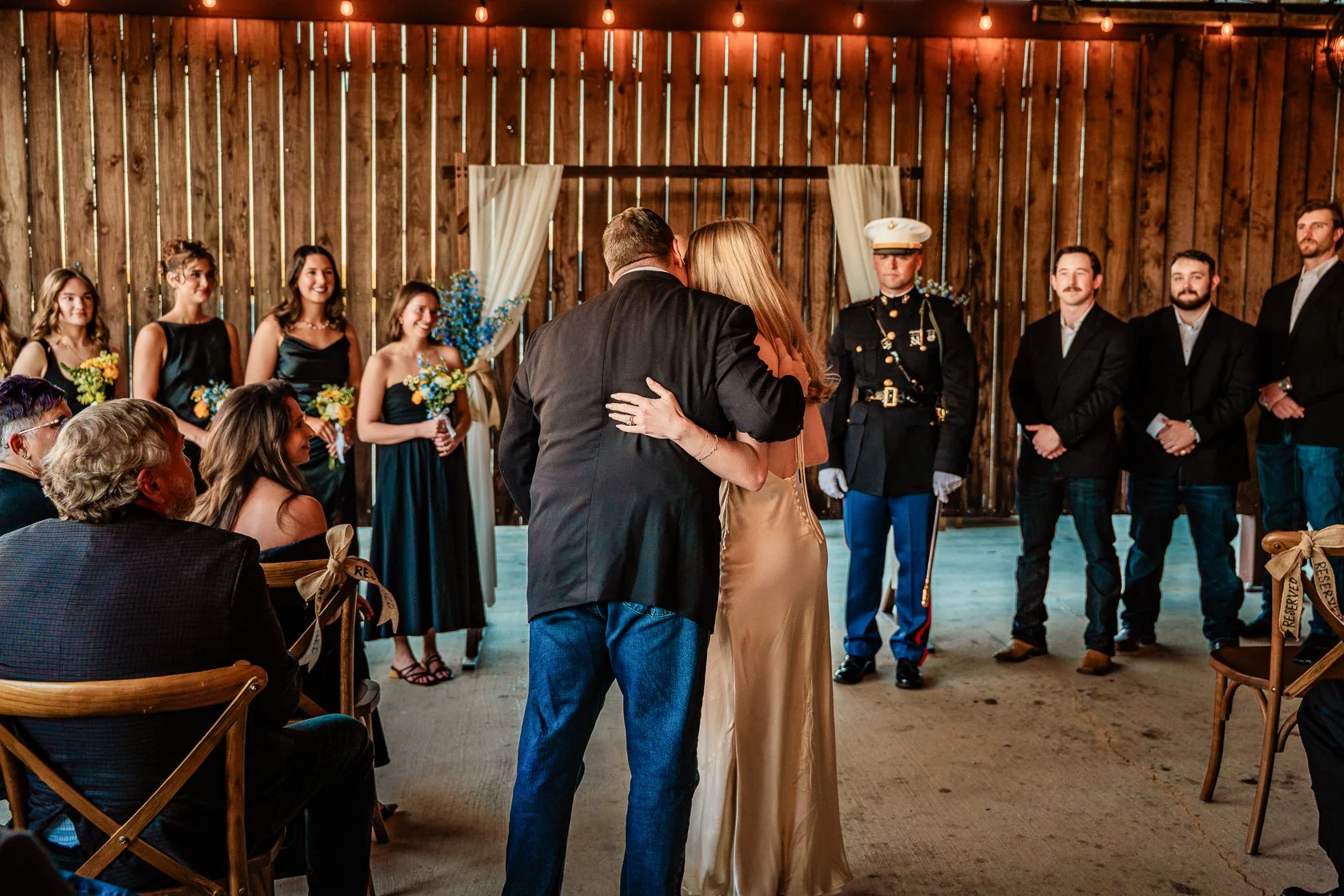 A wedding ceremony taking place in a rustic barn, with the bride and groom sharing a dance in front of seated guests and bridesmaids holding bouquets of yellow and blue flowers, while a uniformed Marine stands at attention nearby.