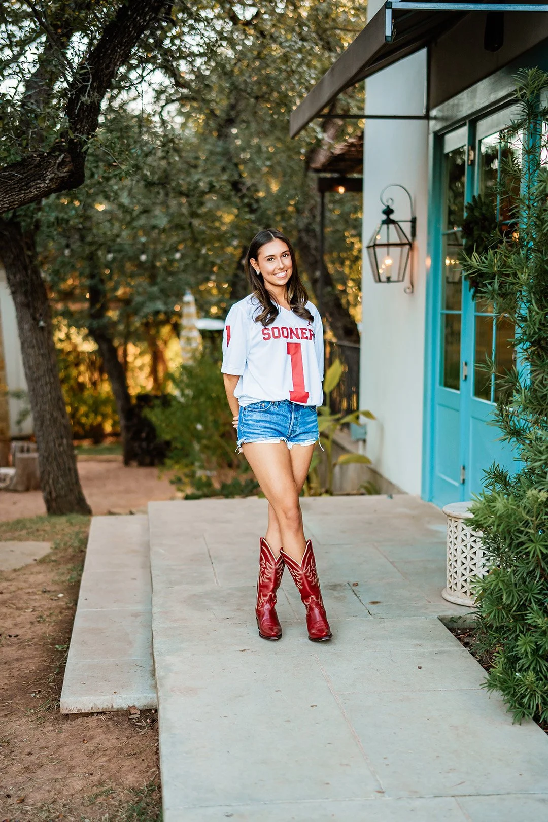 A young woman with long dark hair and a bright smile standing on a sidewalk. She is wearing a white football jersey with 'Sooners 1' on it, denim shorts, and red cowboy boots. The background features a white building with blue doors, teal window fram