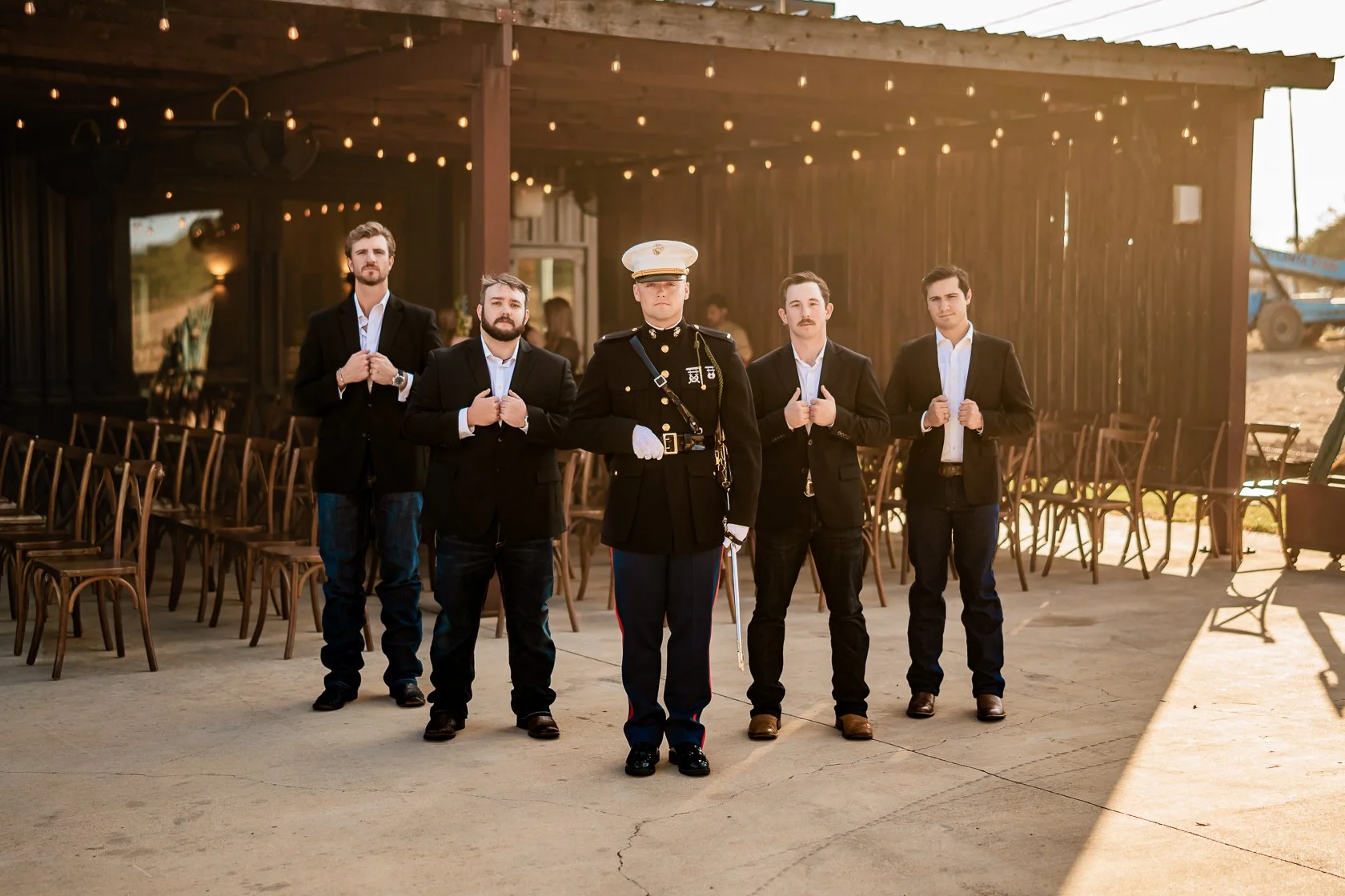 A group of five men, including a military officer, standing in front of a rustic barn with string lights, with chairs behind them and sunlight illuminating the scene.
