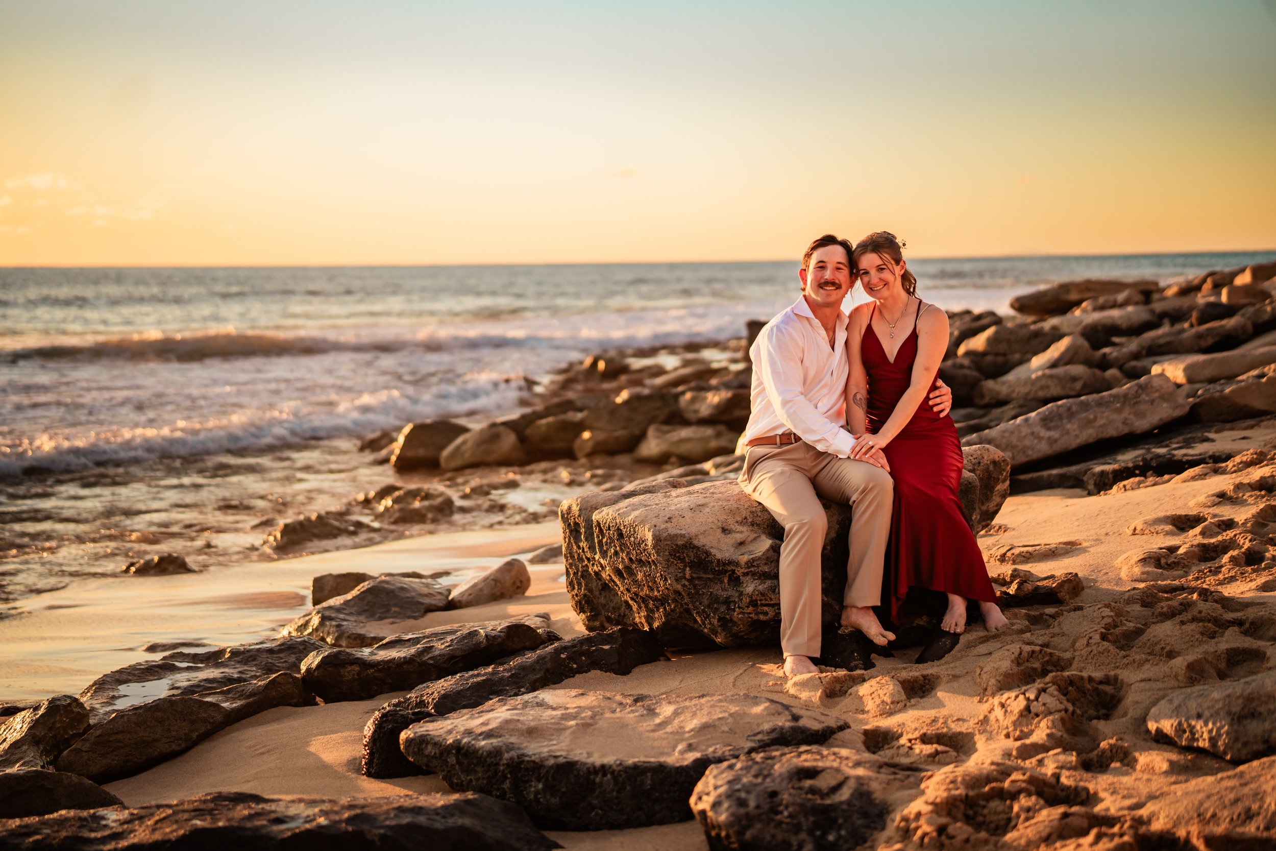 A couple sitting on a large rock on a beach at sunset, near the ocean, smiling and embracing each other.