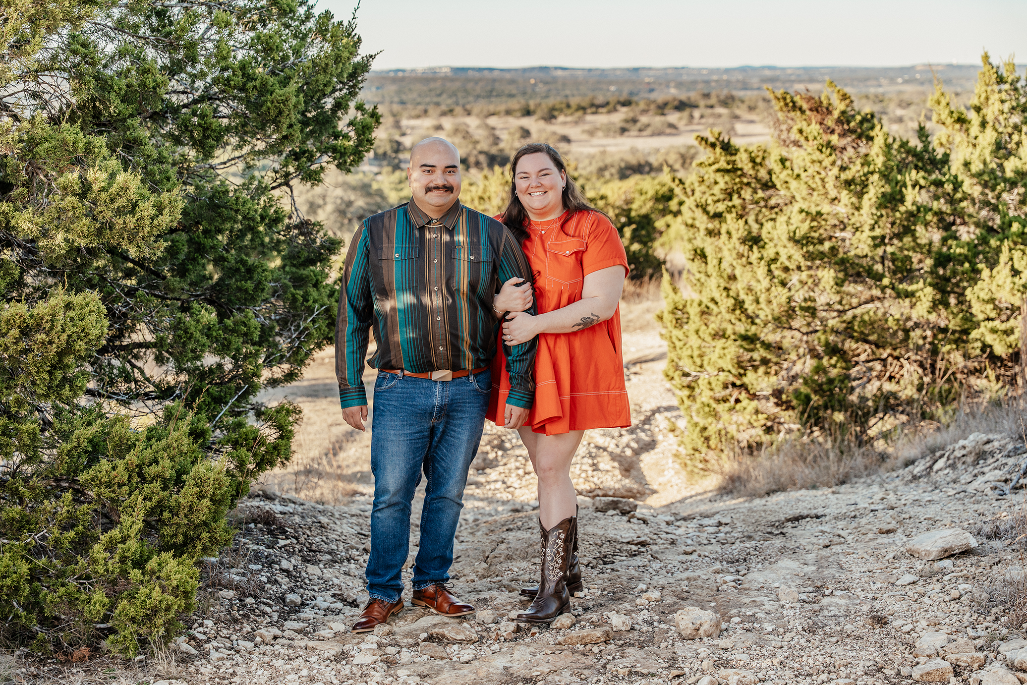 A man and woman standing on a rocky trail in a semi-arid landscape with bushes and trees. The man is wearing a striped button-up shirt and jeans, and the woman is wearing a bright orange dress and cowboy boots. They are smiling and holding onto each 