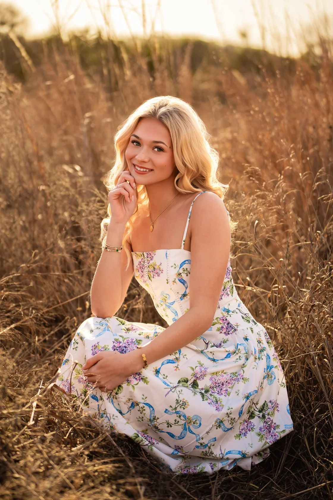 A smiling young woman with blonde hair wearing a floral dress, sitting on dry grass in a field during sunset.