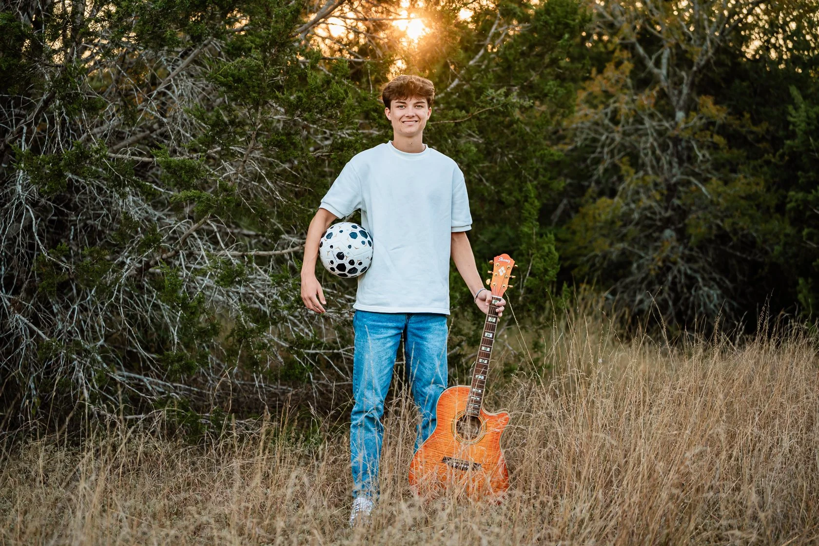 A teenage boy standing outdoors in a grassy field with trees in the background, holding a soccer ball under his left arm and a guitar in his right hand, with the setting sun behind him.