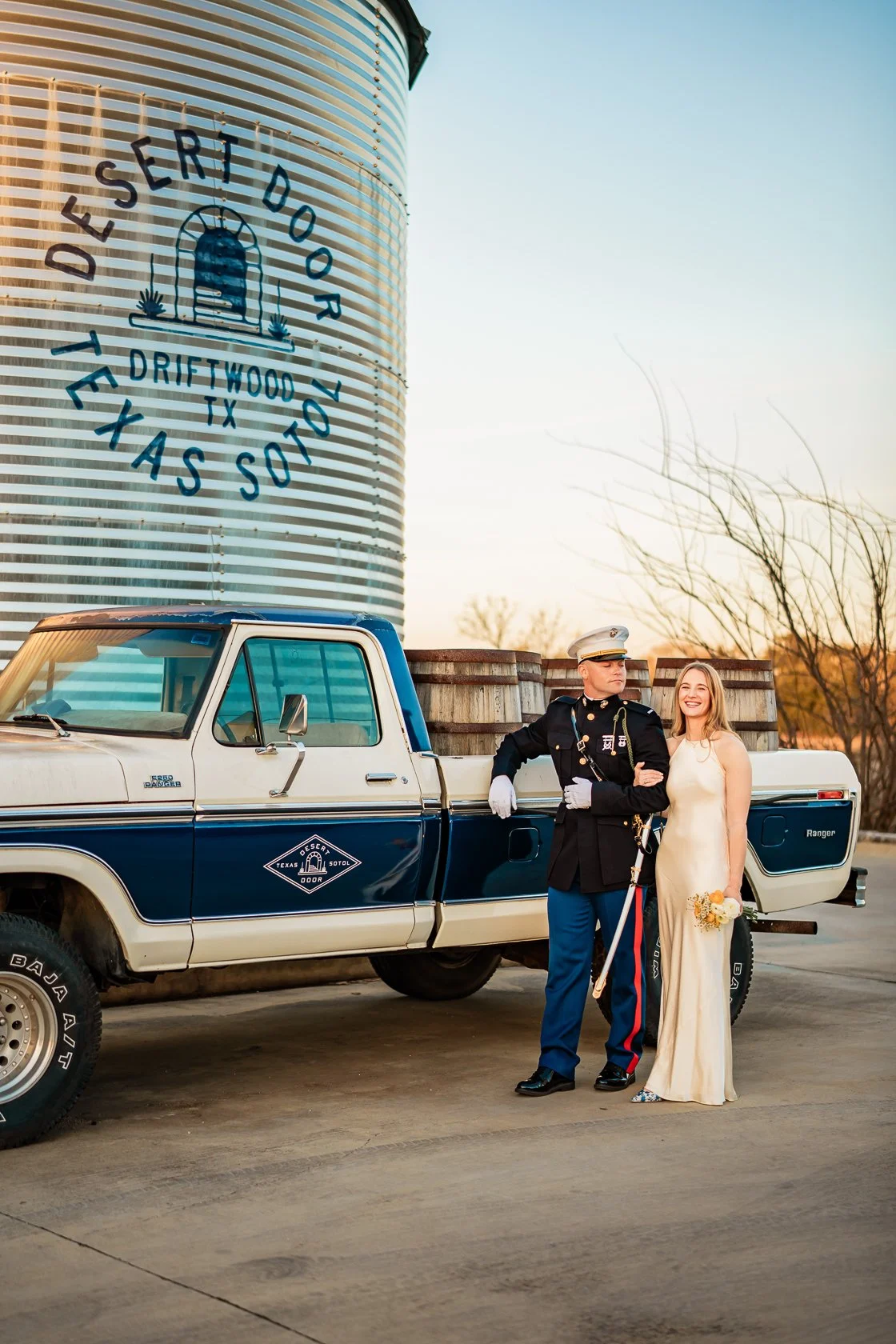A couple dressed in wedding attire standing next to a police officer in uniform, with a vintage pickup truck in the background, in front of a large agricultural silo with the sign "Desert Door: The Texas Sotol, Driftwood TX."