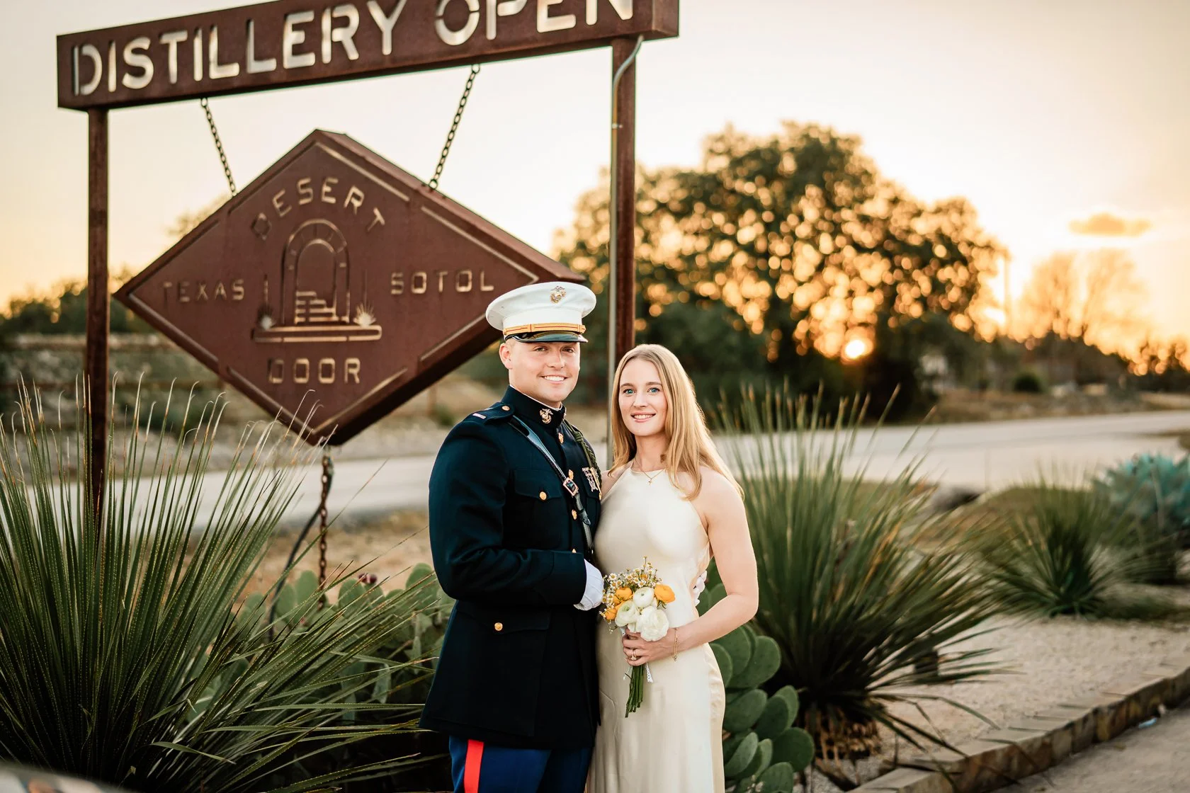 A couple dressed in wedding attire poses for a photo in front of a sign that says 'Distillery Open' at dusk. The groom wears a military uniform and a white cap, and the bride wears a white dress holding a bouquet of white and yellow flowers. There ar