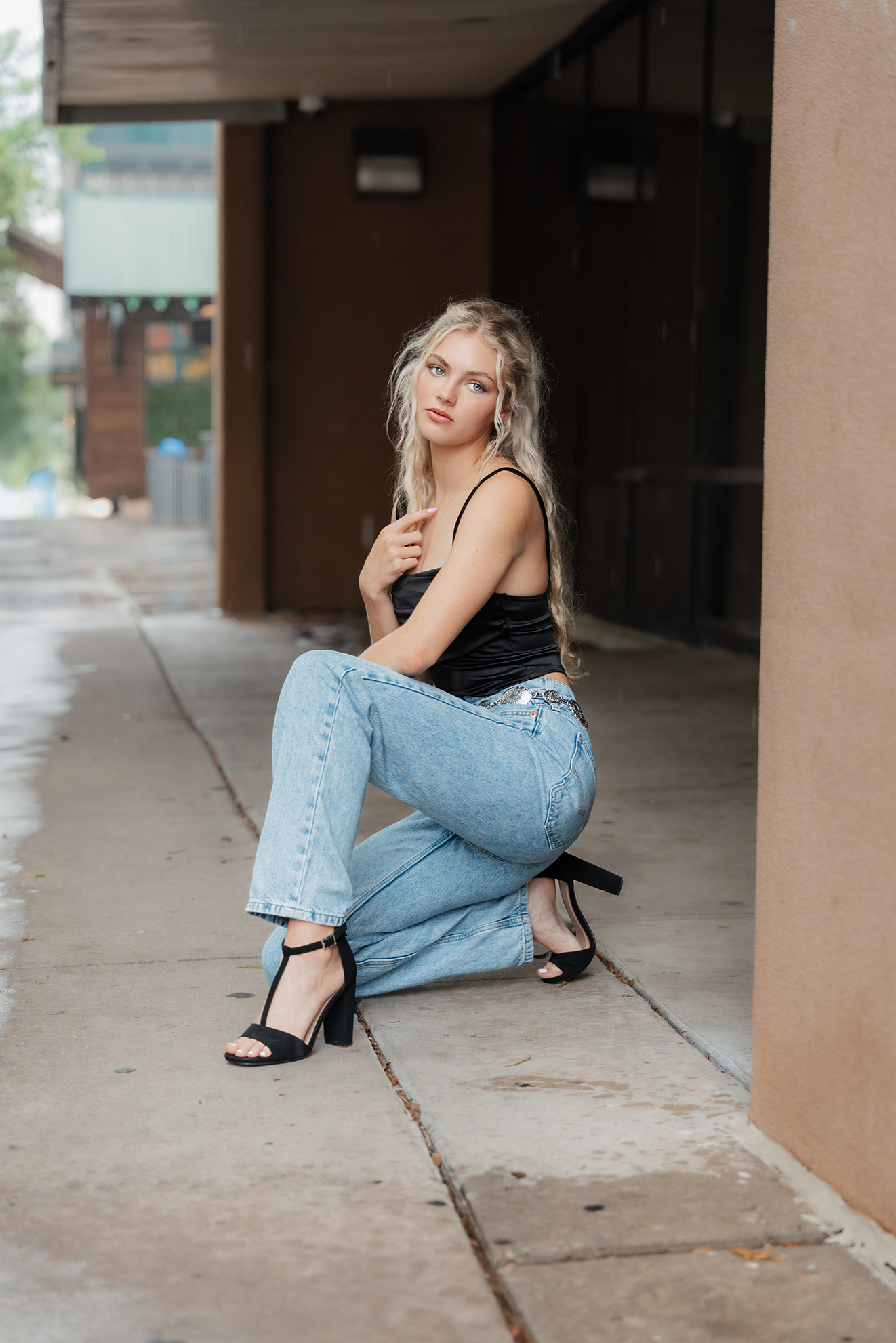 A young woman with wavy blonde hair, wearing a black tank top, blue jeans, and black high heels, kneeling on a sidewalk in an urban area.