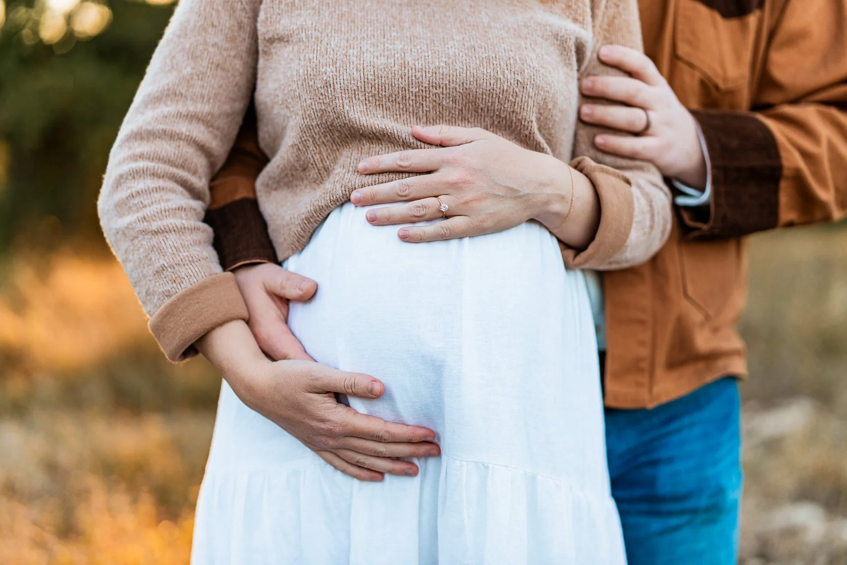 A pregnant woman in a light blue dress is being gently held by a partner, with hands on her belly, outdoors during fall with blurred background.