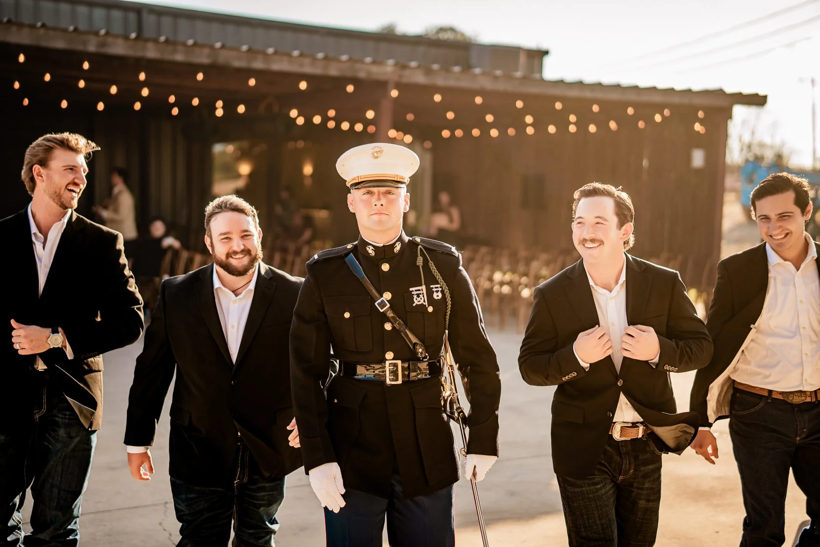 A group of men in suits and a military officer in uniform are standing outdoors, smiling and laughing with a barn decorated with string lights in the background.