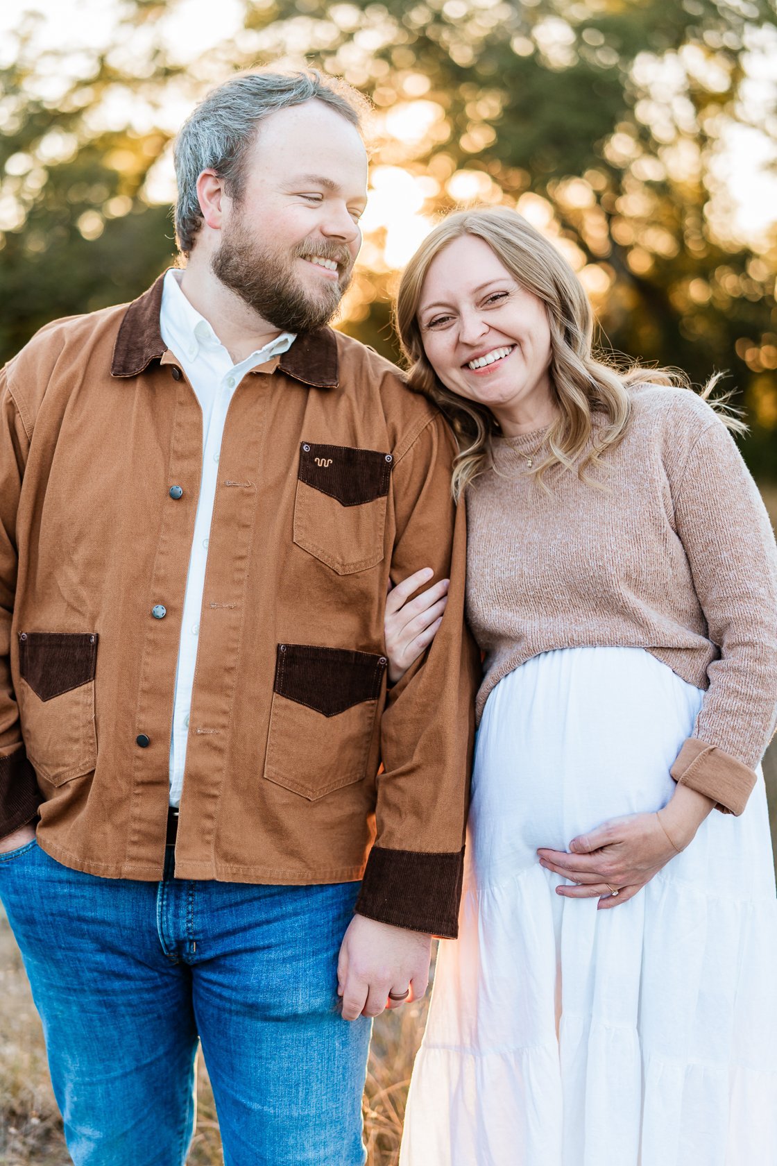 A smiling pregnant woman with her hand on her belly, standing next to a happy man outdoors at sunset.