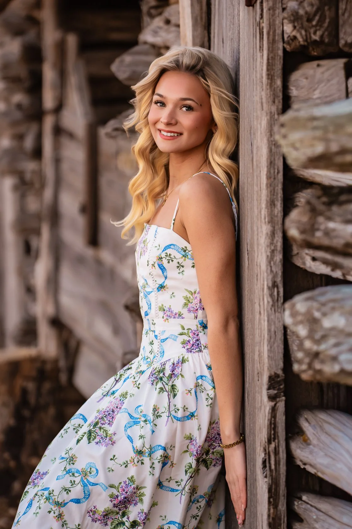 A young woman with long blonde hair, smiling, leaning against a wooden wall outdoors, wearing a white floral sundress with spaghetti straps.