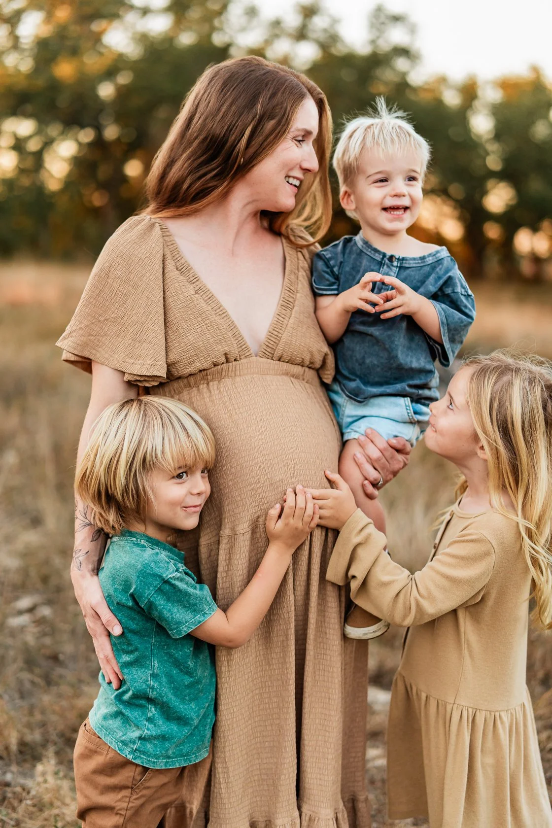 A pregnant woman smiling and holding a toddler, surrounded by her two young children, outdoors during sunset.