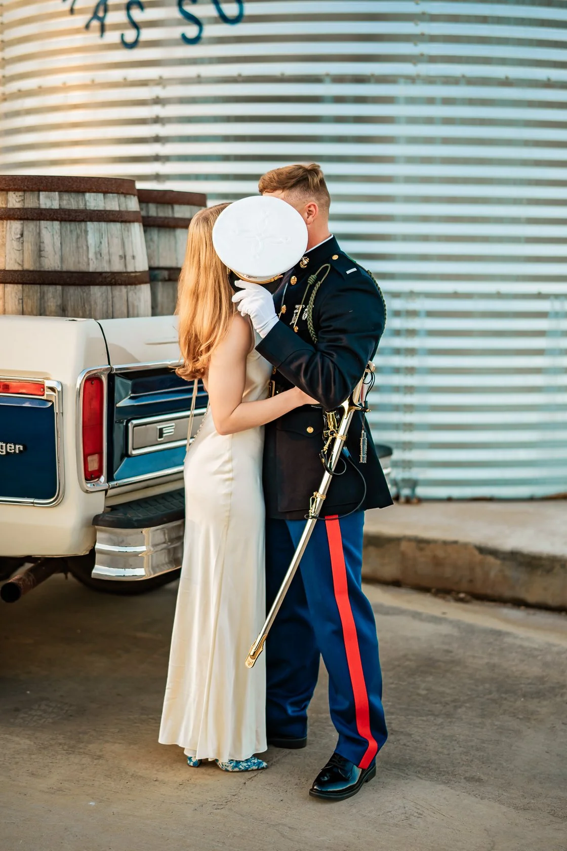 A woman and a soldier are kissing in front of a vintage truck. The woman is wearing a white dress, and the soldier is in a formal military uniform. The woman covers her face with a round white object as they embrace.