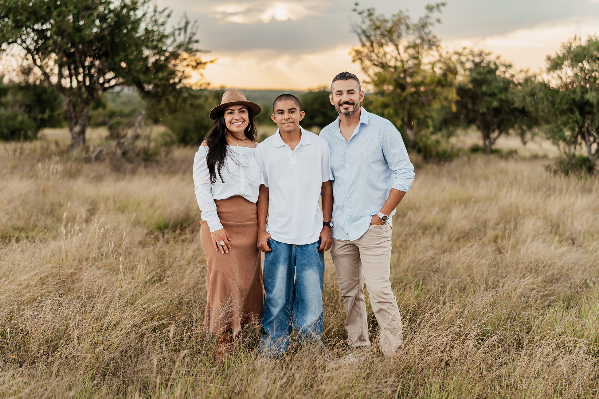 Family of three standing in a grassy field with trees and a cloudy sky in the background during sunset.