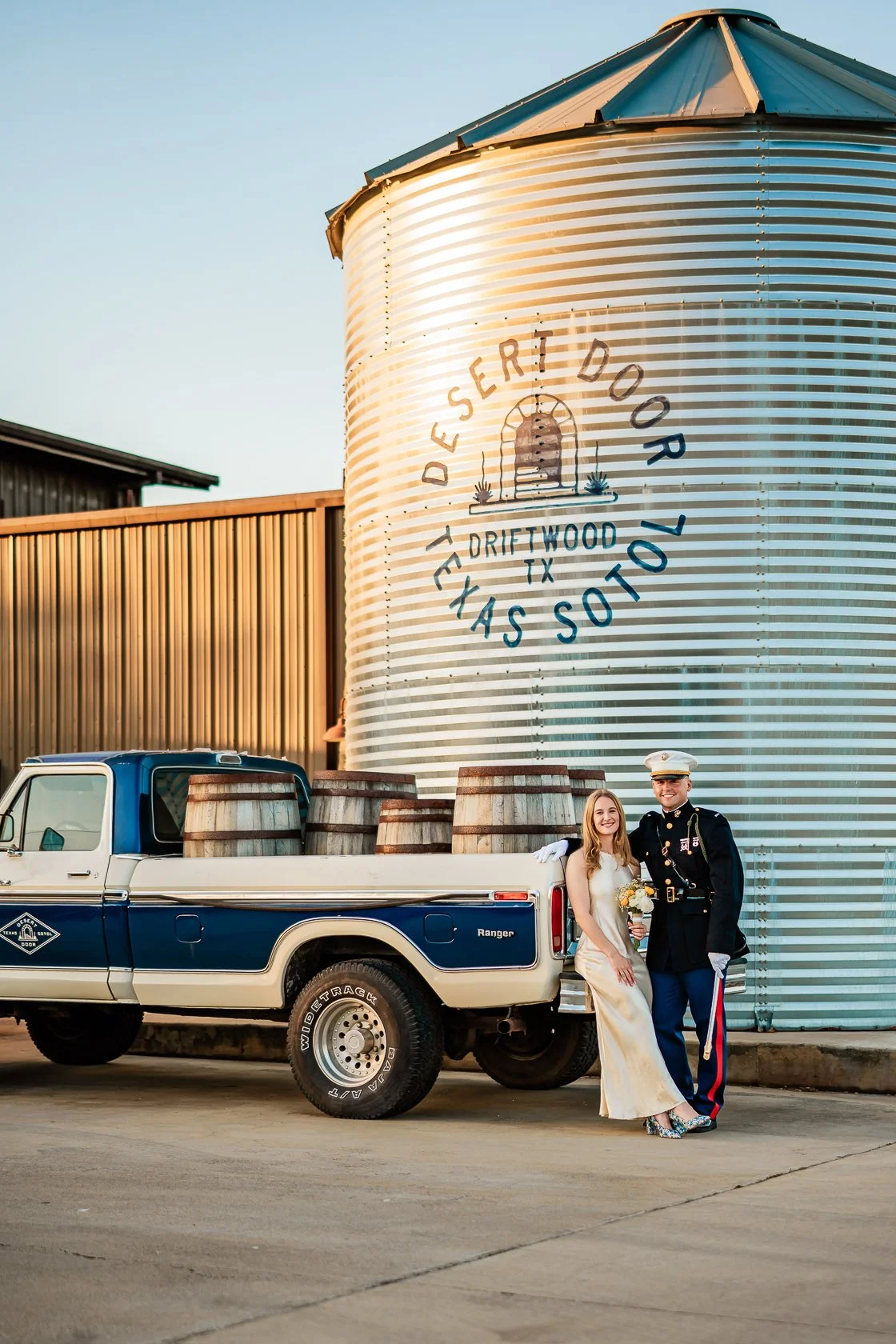 A bride and groom in wedding attire standing next to a truck filled with barrels, in front of a large silos with the words "Delivered Door Thru!, Drietwood TX, Has So!," painted on it.