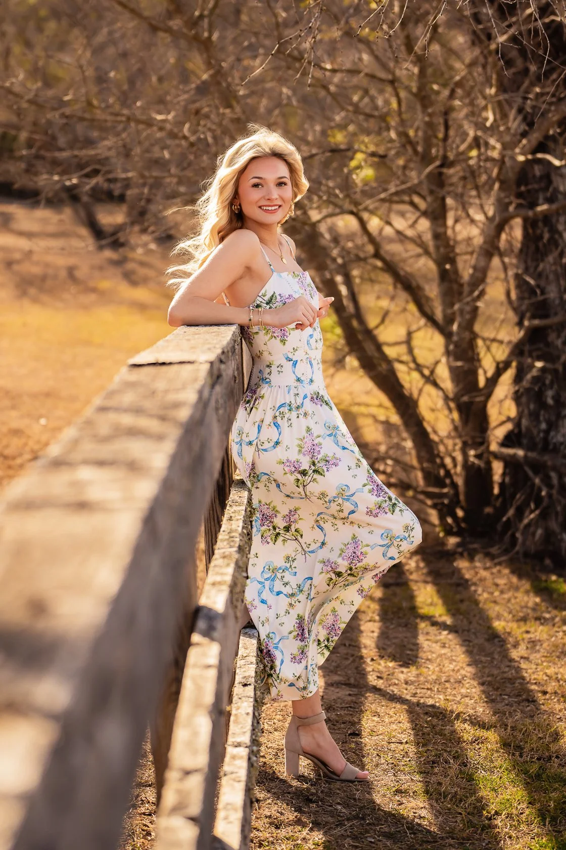 A young woman in a floral dress leans on a wooden fence in an outdoor setting with bare trees and autumn-colored ground.