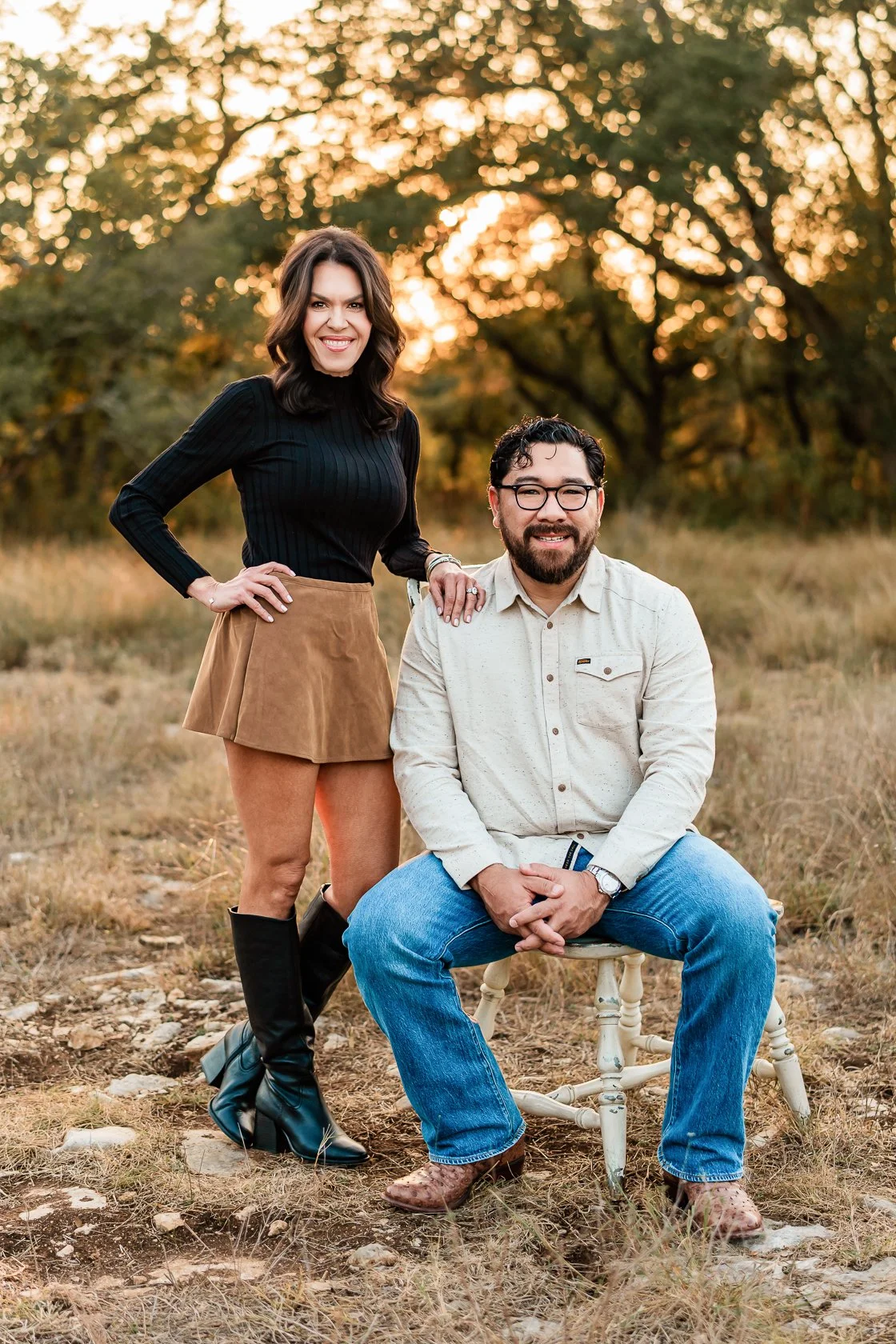 A woman and a man outdoors during sunset. The woman is standing with one hand on her hip, wearing a black long-sleeve top, a tan skirt, and black knee-high boots. The man is seated on a wooden chair, wearing a light-colored button-up shirt, jeans, an