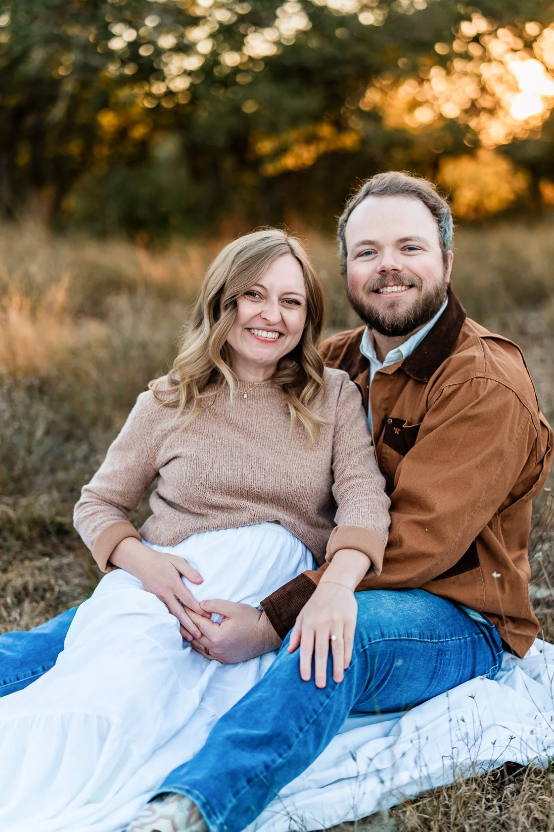 A smiling pregnant woman with blonde hair in a beige sweater sitting on a blanket outdoors, holding her belly, next to a smiling man with a beard in a brown jacket, in a field with trees and sunset in the background.